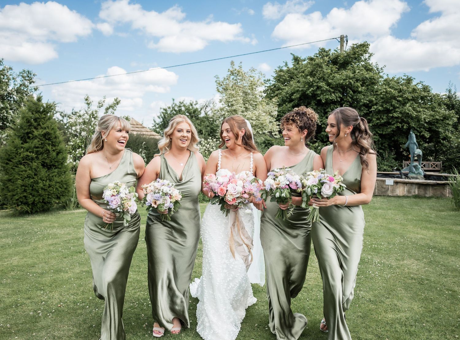Wedding party poses outdoors with bridesmaids in sage green dresses holding white bouquets.