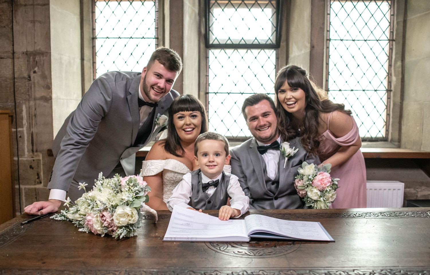 Wedding party gathers around wooden table for document signing in historic room with leaded windows.