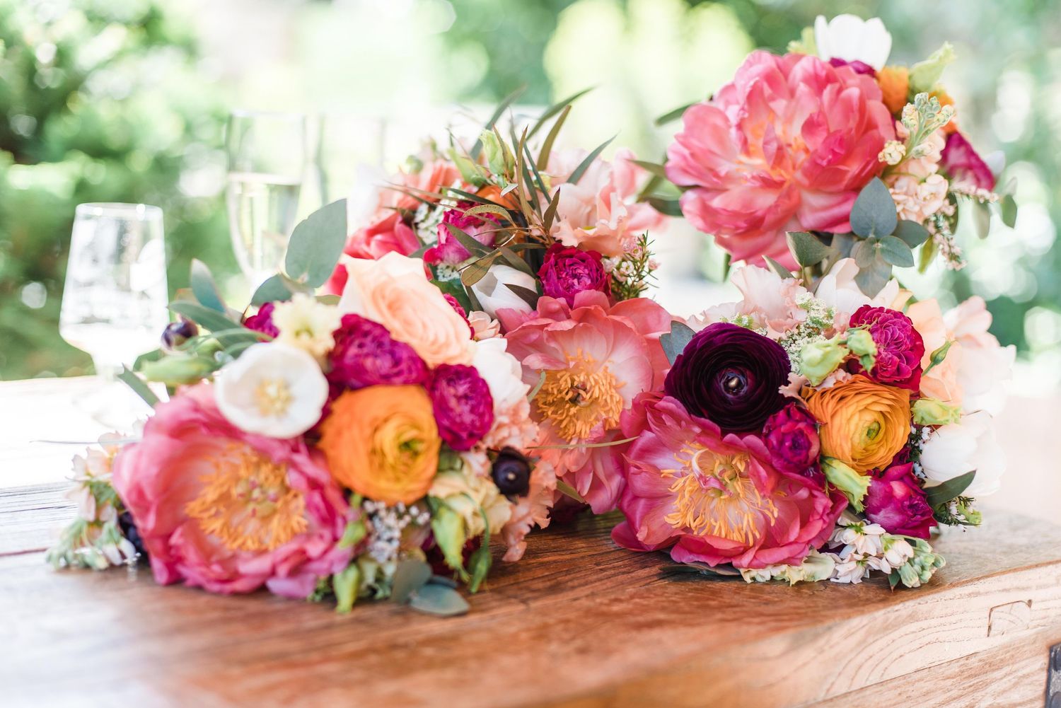 Vibrant pink and coral wedding bouquets with roses and ranunculus arranged on wooden table with wine glass in background.