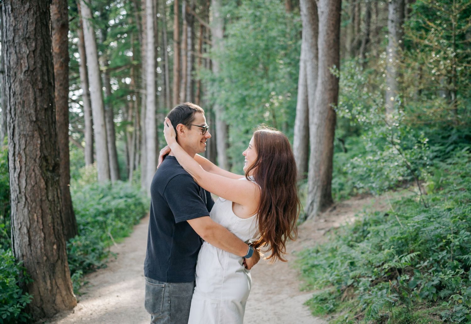 A loving couple embraces on a forest path surrounded by tall evergreen trees and lush greenery.