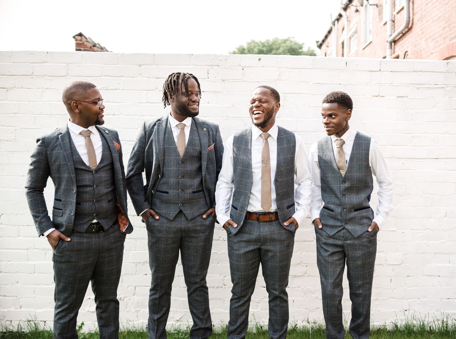 Four men in formal suits and vests pose against a white wall outdoors.