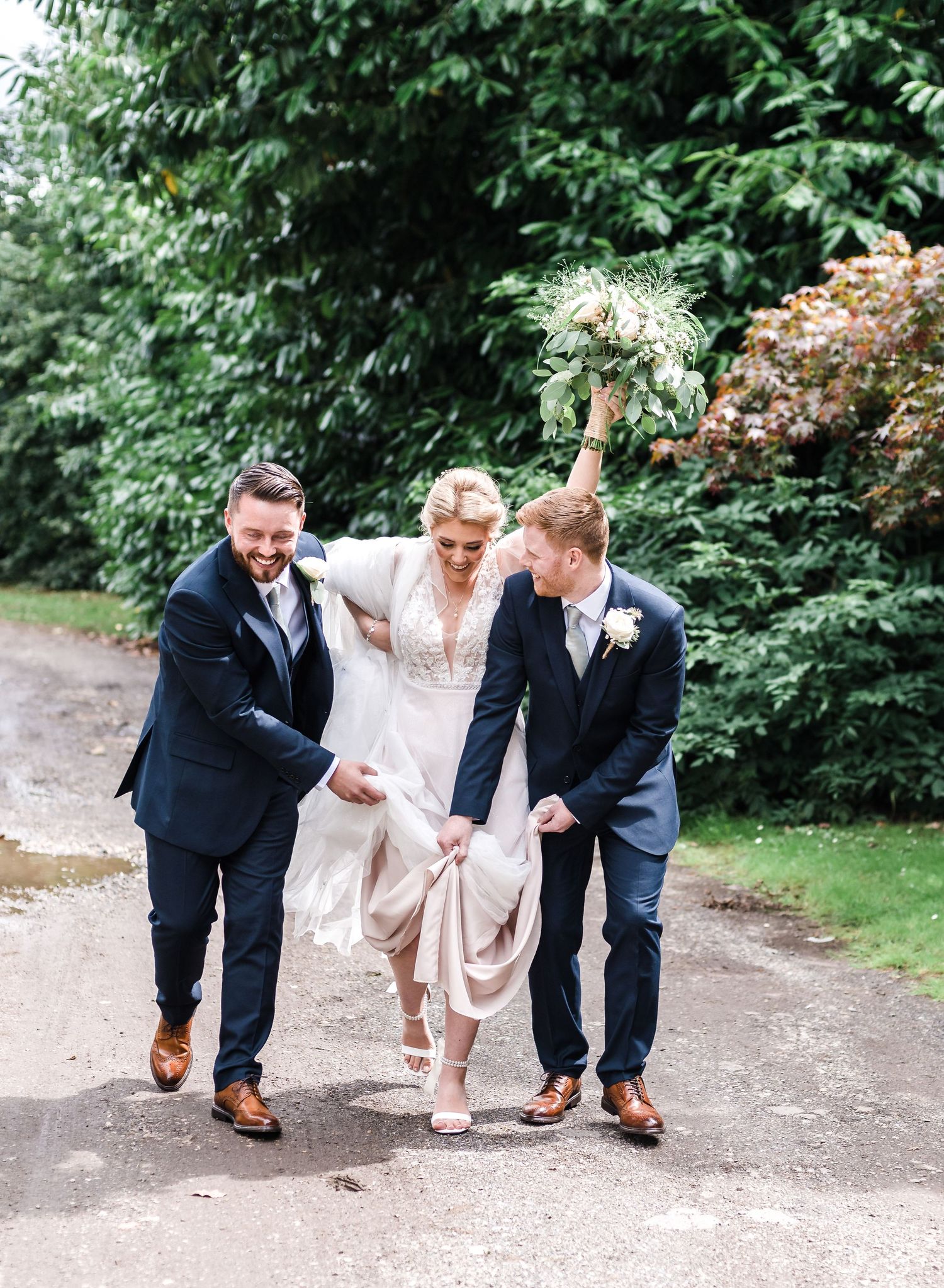 Wedding party walking and laughing together on a tree-lined path while helping with a dress train.