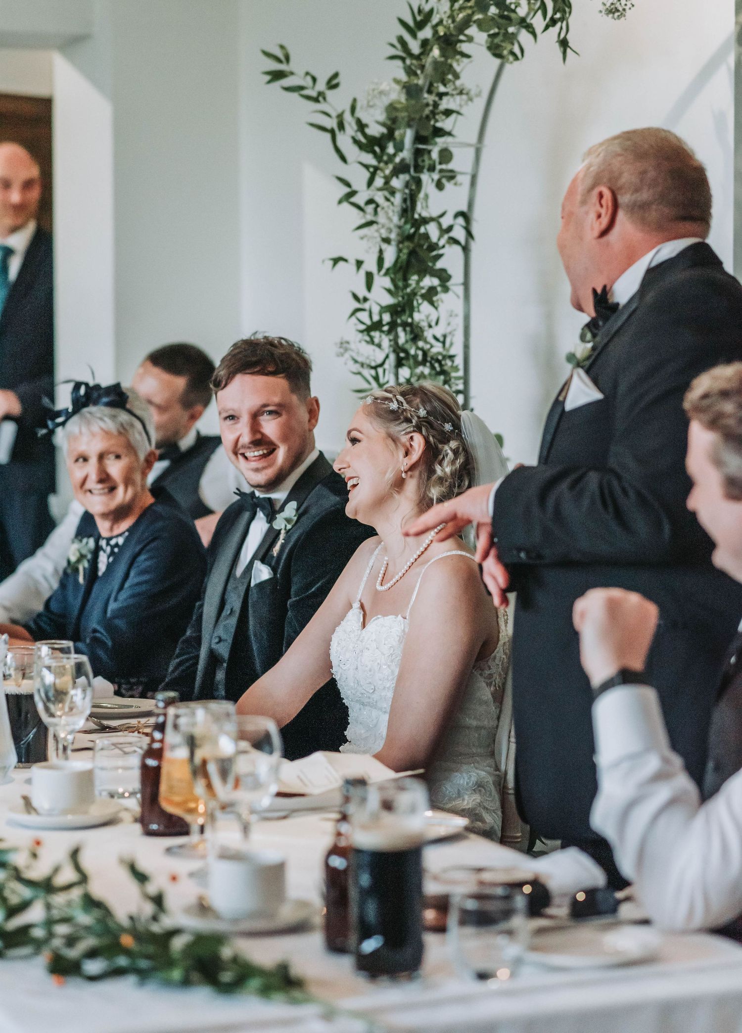 Candid wedding reception moment showing laughter at head table decorated with greenery.