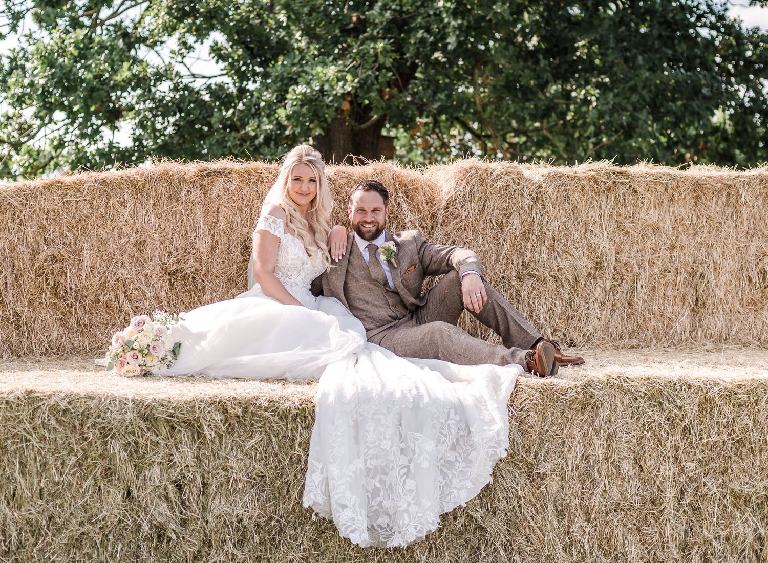 Newlyweds sitting together on large hay bales during outdoor countryside wedding.