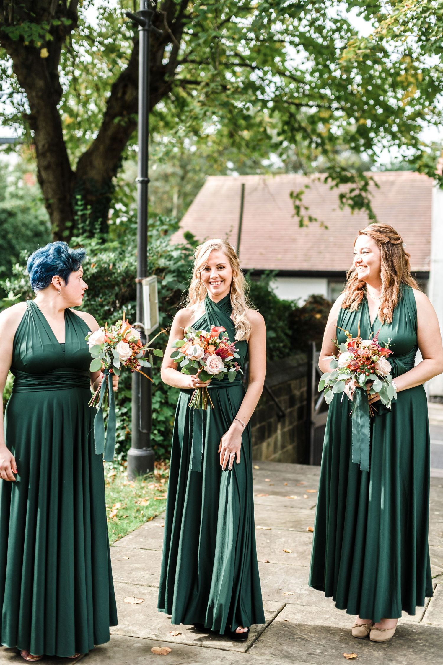 Bridesmaids in floor-length green dresses holding floral bouquets outdoors.