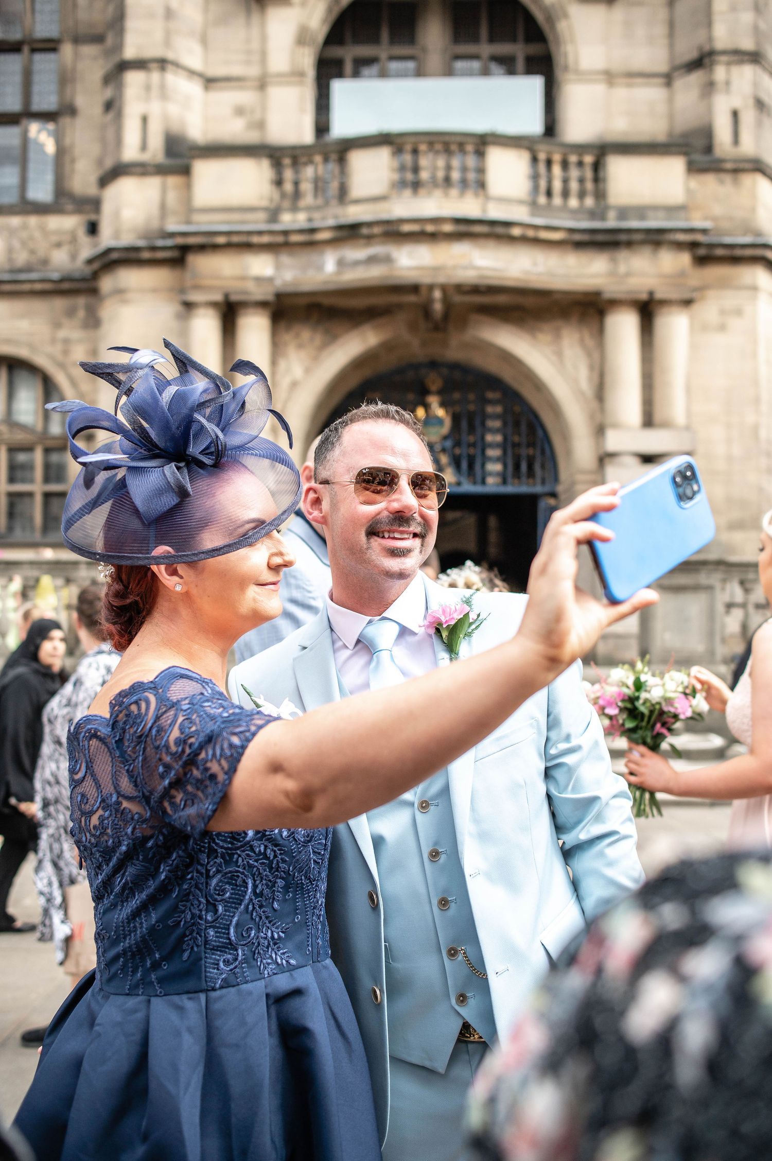Guest in blue dress and fascinator taking selfie outside historic building during wedding celebration.