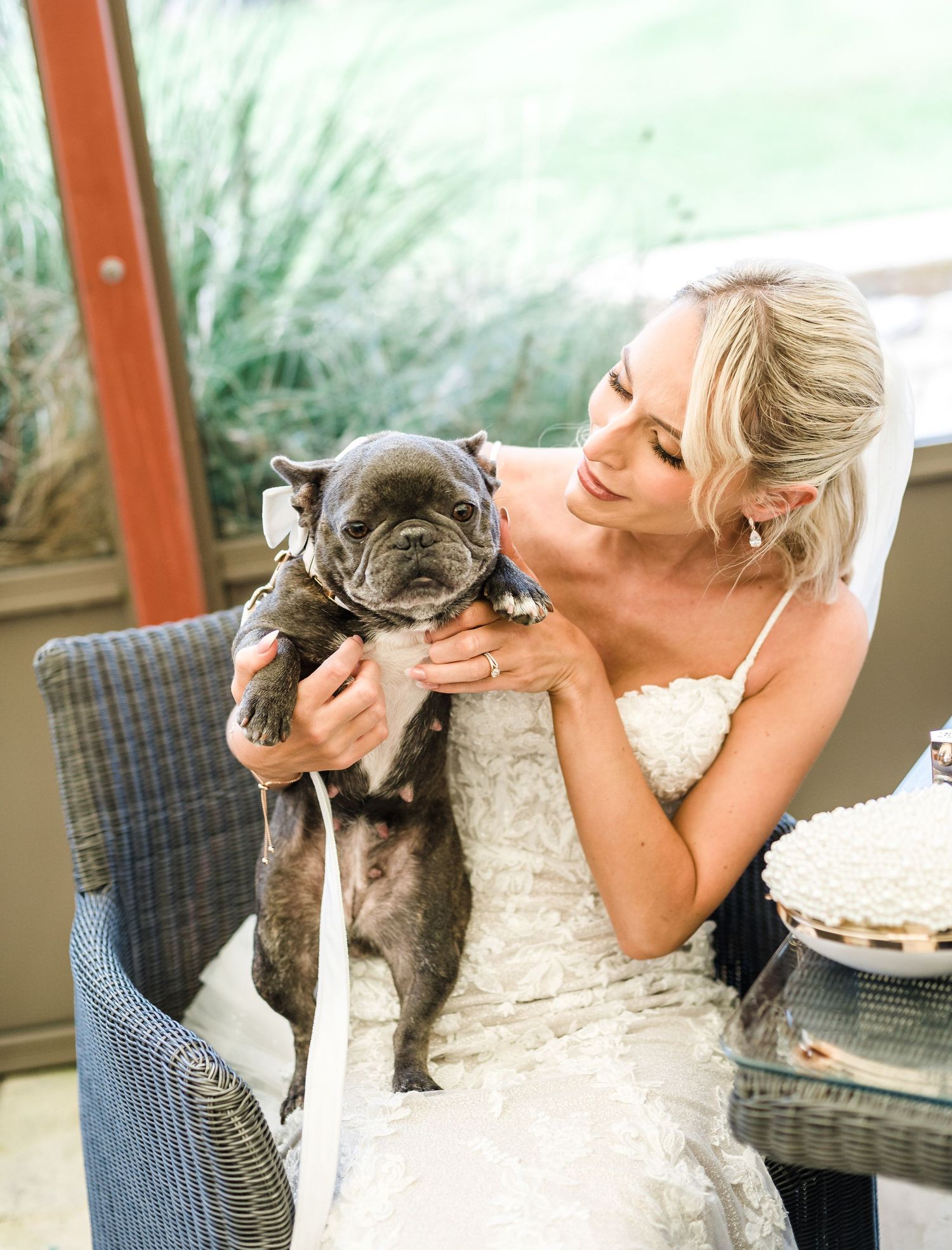 Bride in white wedding dress cuddles with grey French Bulldog in outdoor setting.