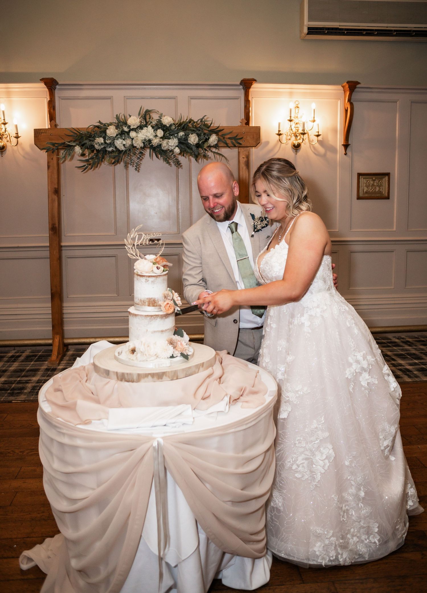 A couple cuts their elegant three-tier white wedding cake beneath a wooden arch with floral decorations.