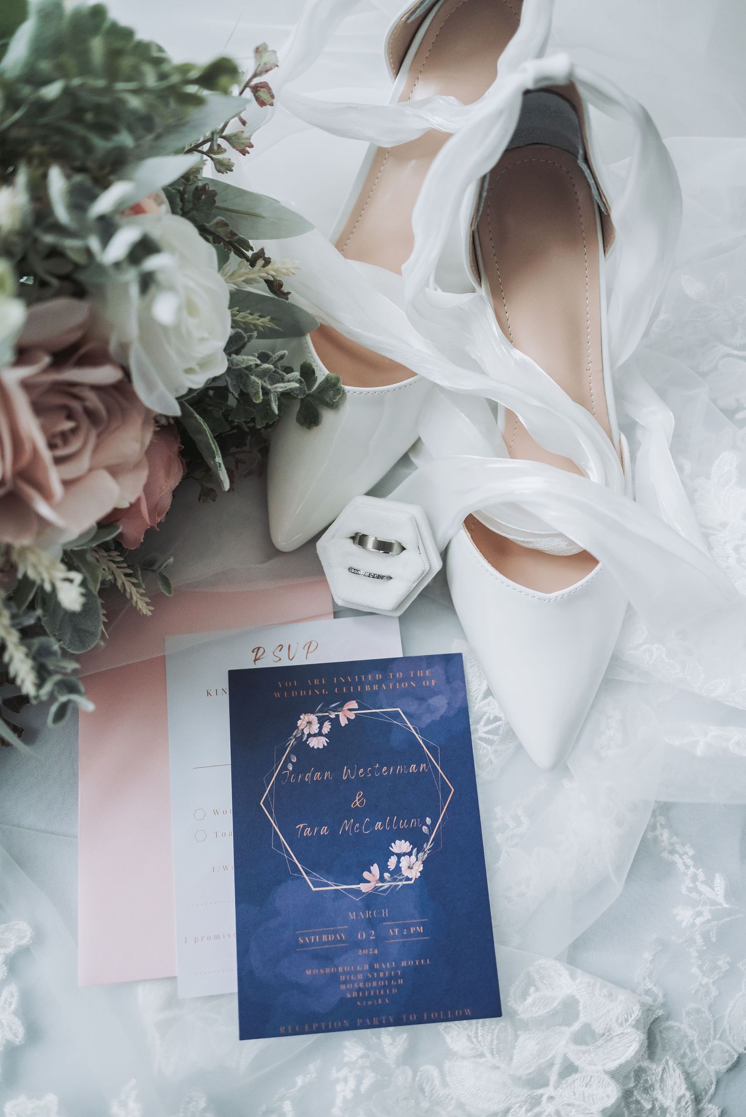 Wedding details featuring white shoes, a navy blue invitation, and pink roses.