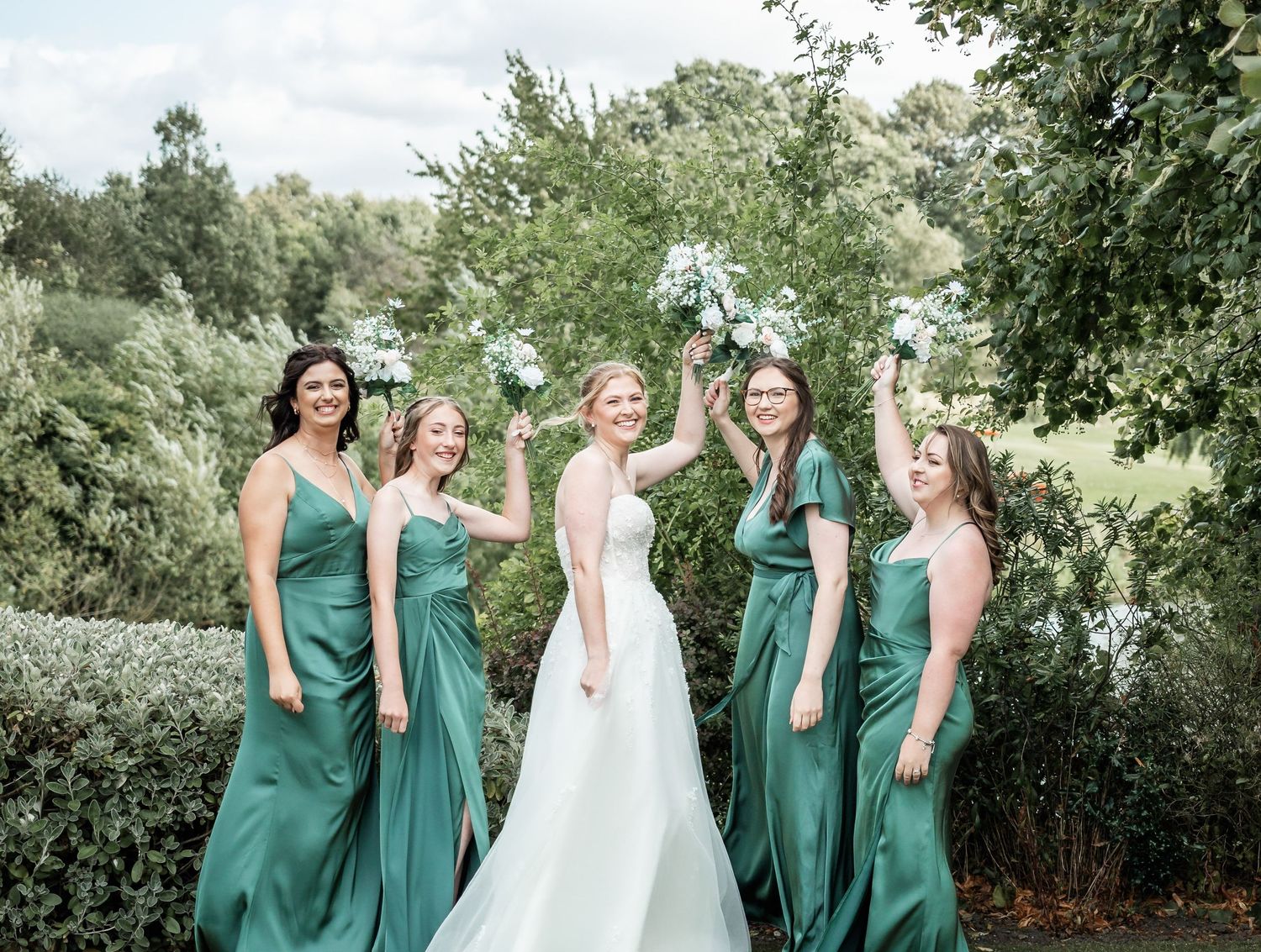 Wedding party poses outdoors wearing elegant emerald green dresses against natural greenery.