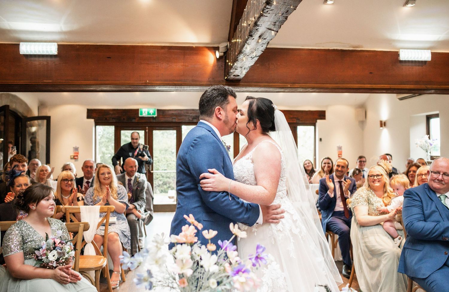 Newlyweds share their first dance while guests applaud in a rustic venue with wooden beams.