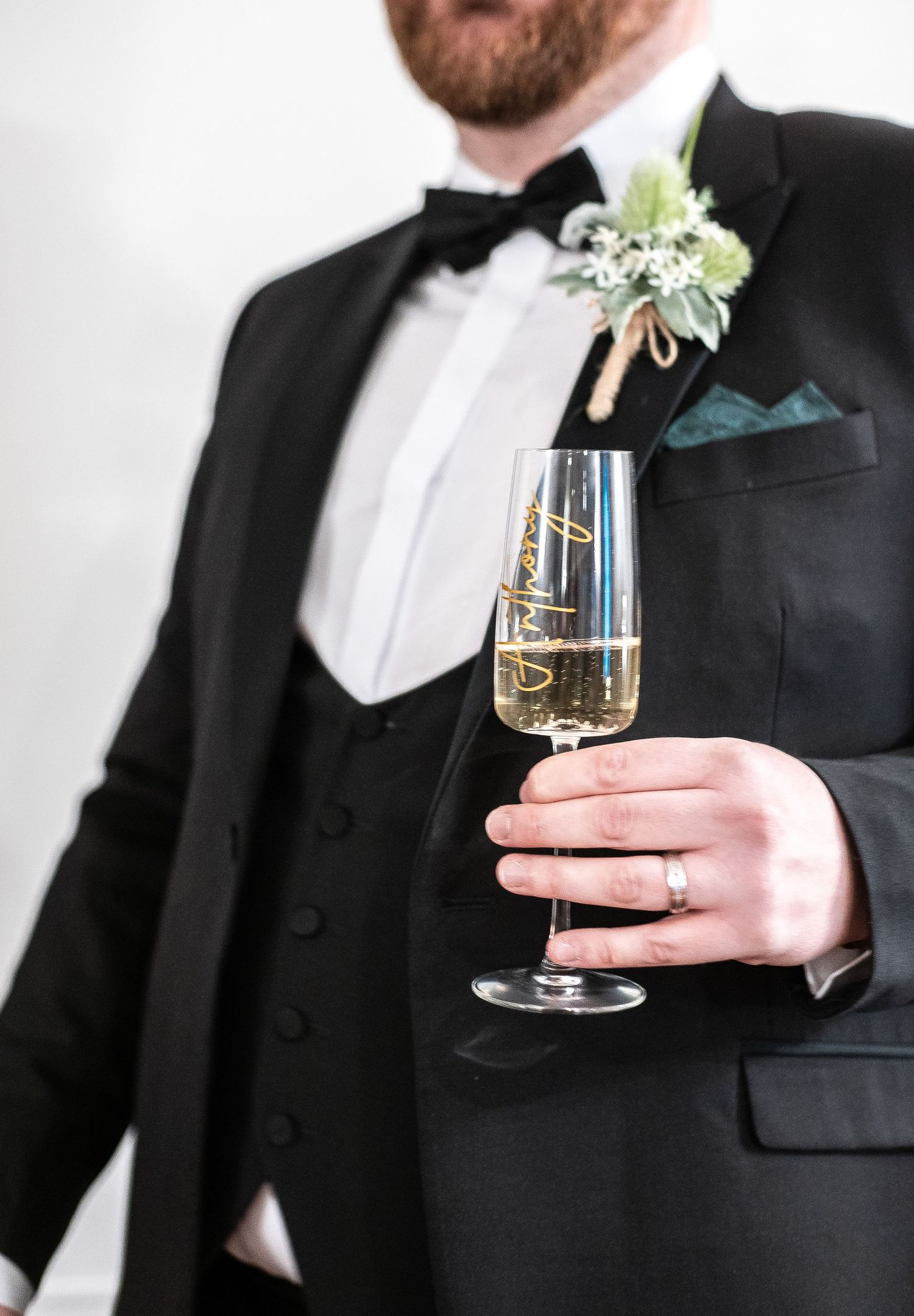 Groom in tuxedo holding champagne glass with boutonniere and teal pocket square detail.