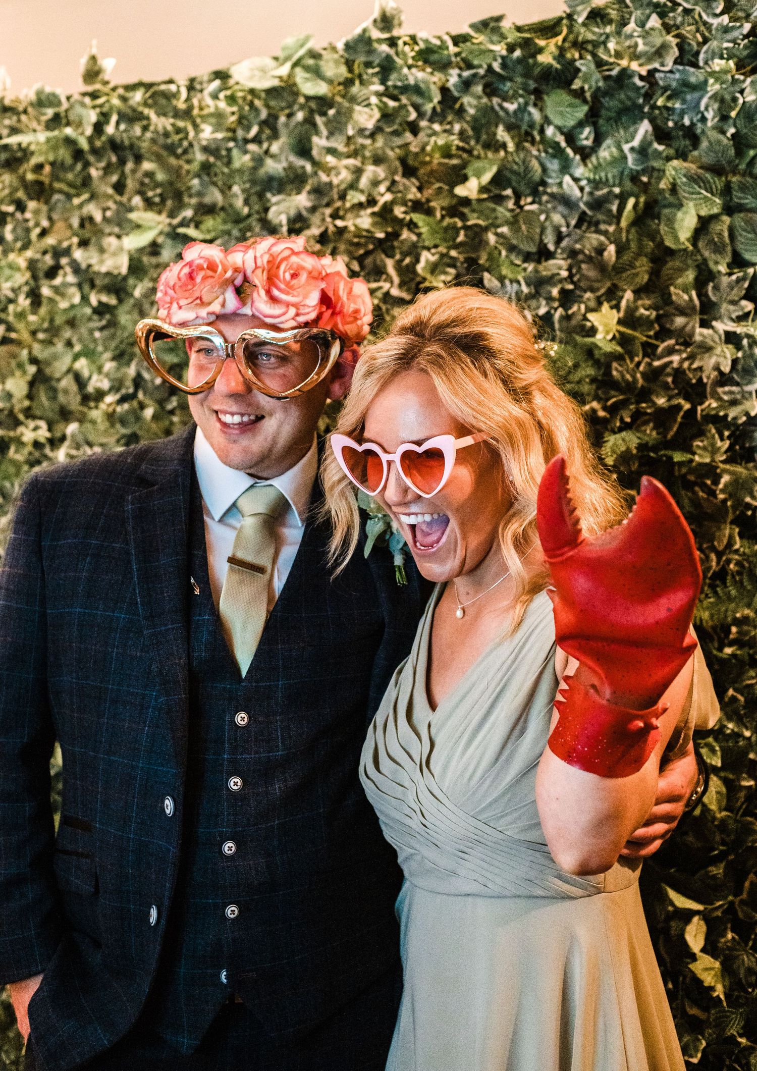 Couple wearing fun sunglasses while posing against greenery backdrop at wedding celebration.