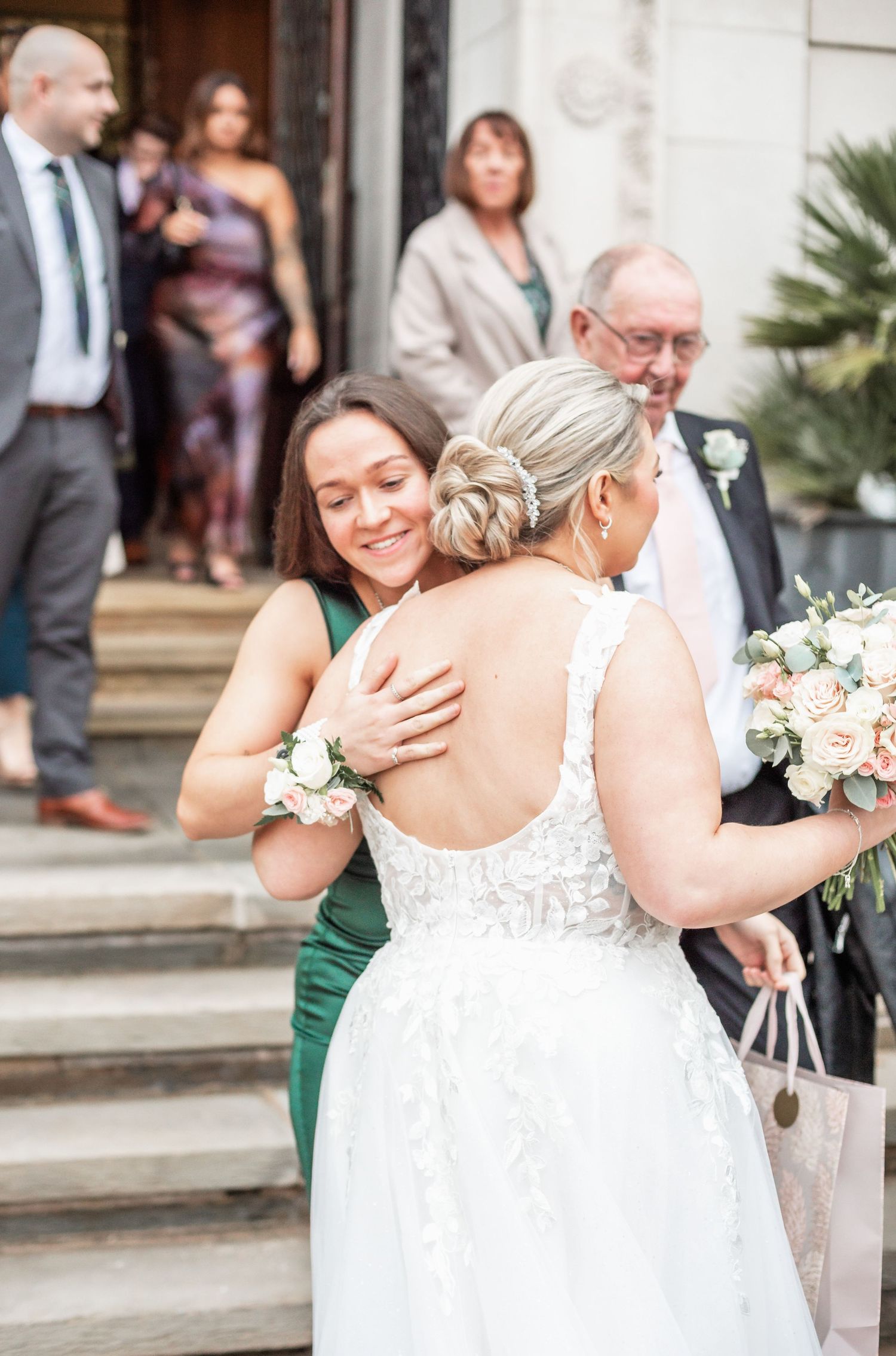 Wedding guests embrace on stone steps outside a building with decorative palm plants.