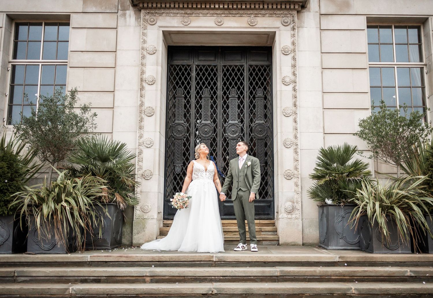 Couple stands on stone steps in wedding attire in front of historic building entrance.