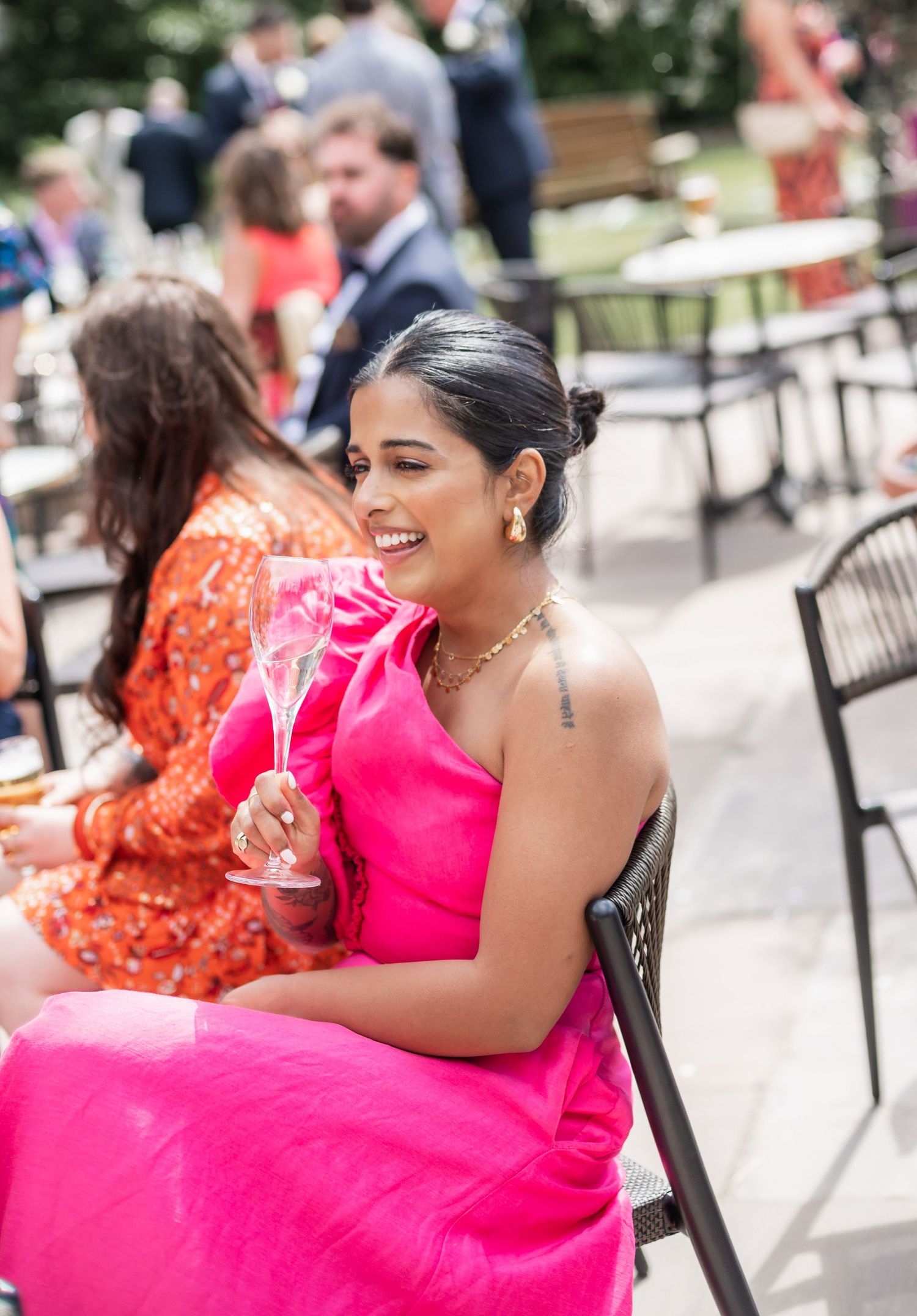 Woman in vibrant pink dress smiles while holding a champagne glass at an outdoor social gathering.