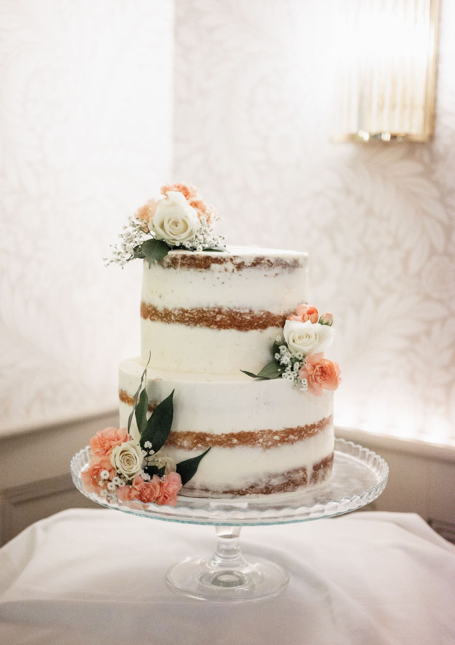 Semi-naked wedding cake decorated with peach and white flowers on crystal cake stand.