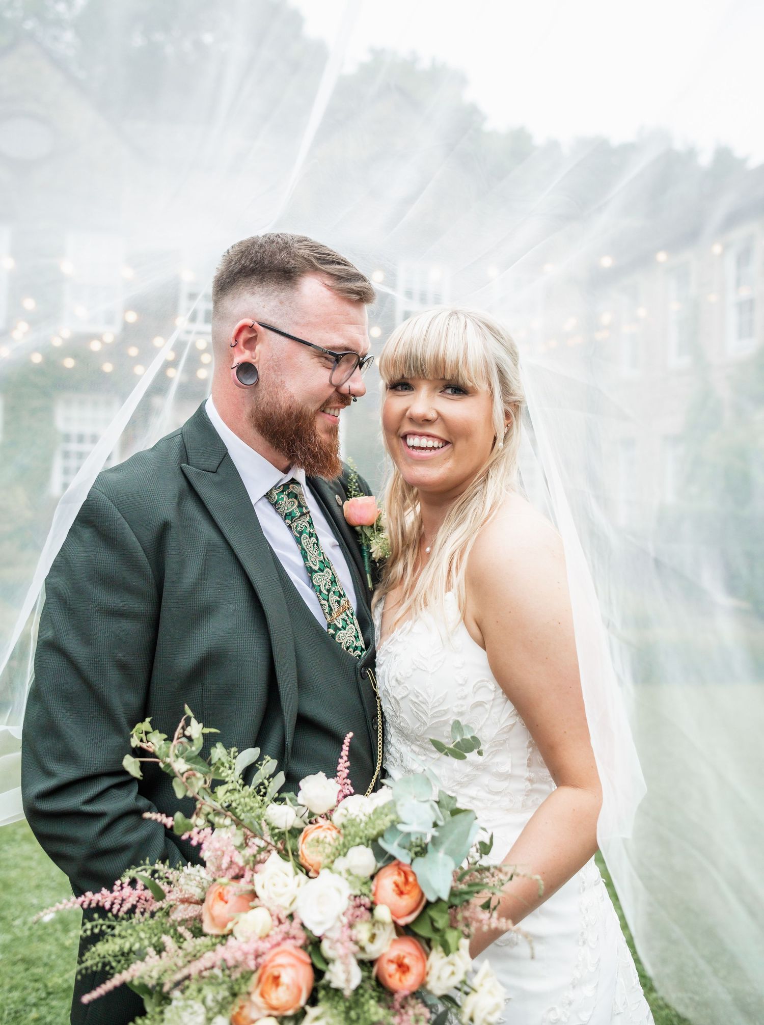 Newlywed couple pose together with a vibrant peach and white bouquet against a dreamy outdoor backdrop with flowing veil.