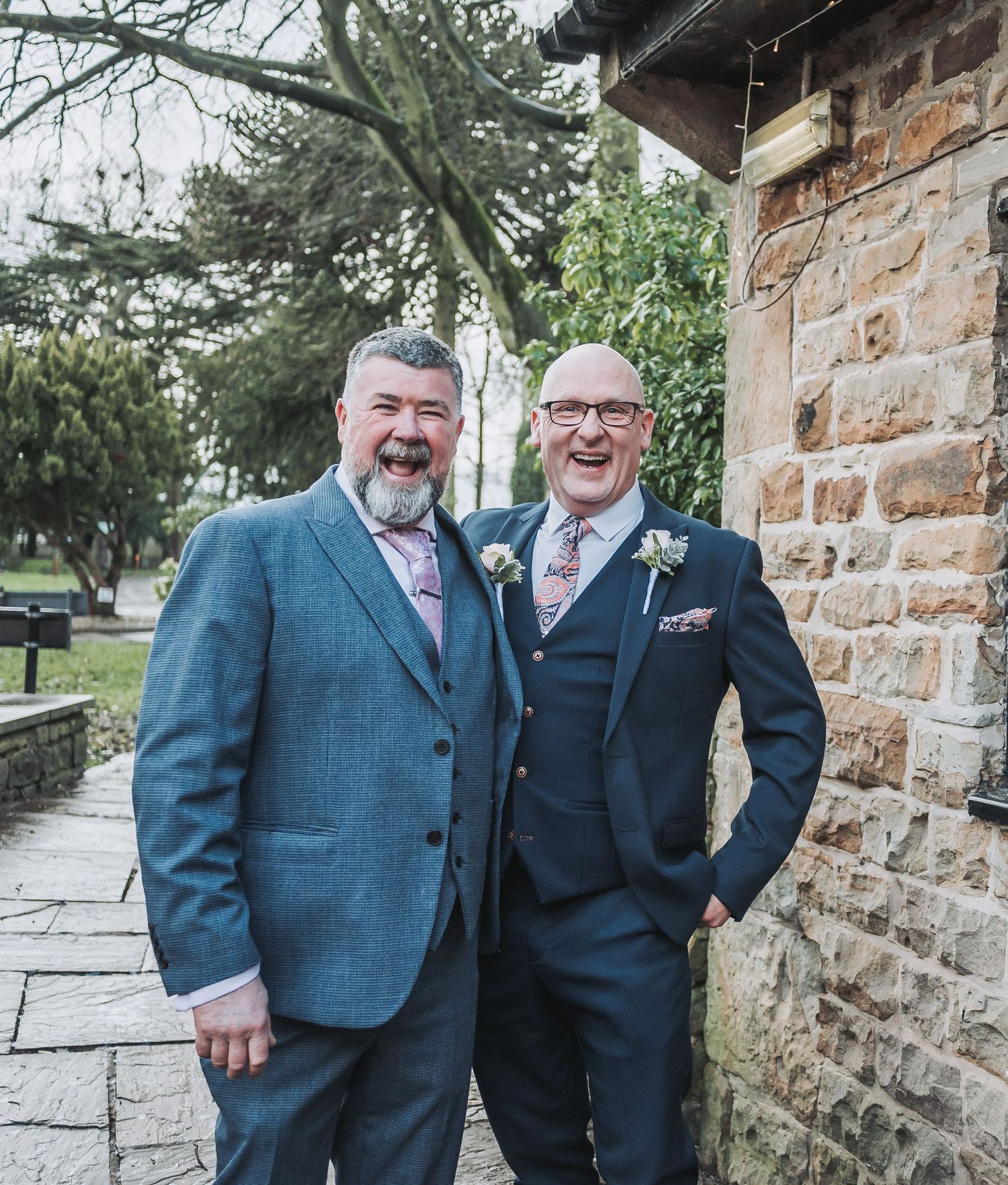 Two guests in blue suits pose together outside a stone building.