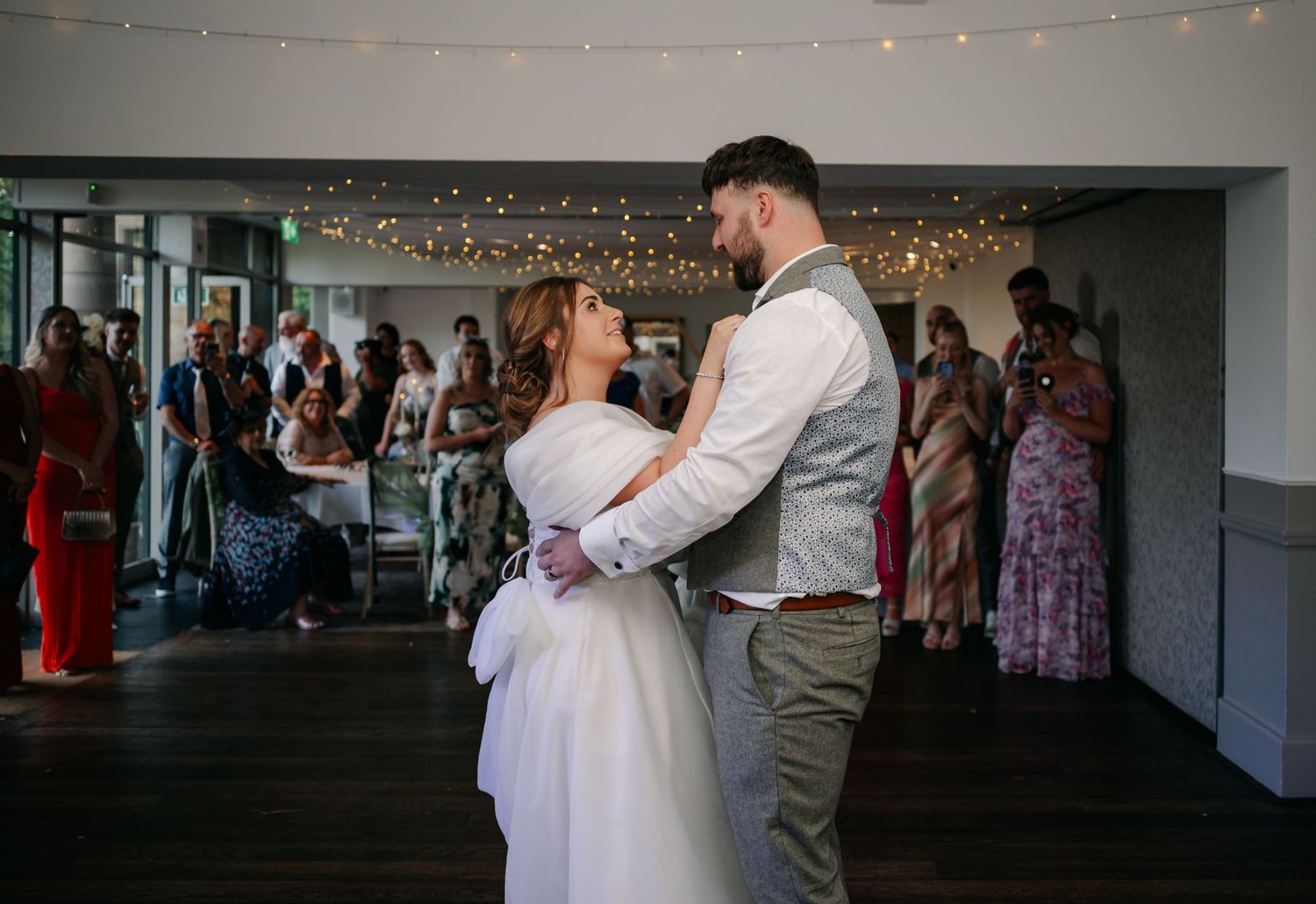 A couple shares their first dance at a wedding reception under string lights with guests watching in the background.