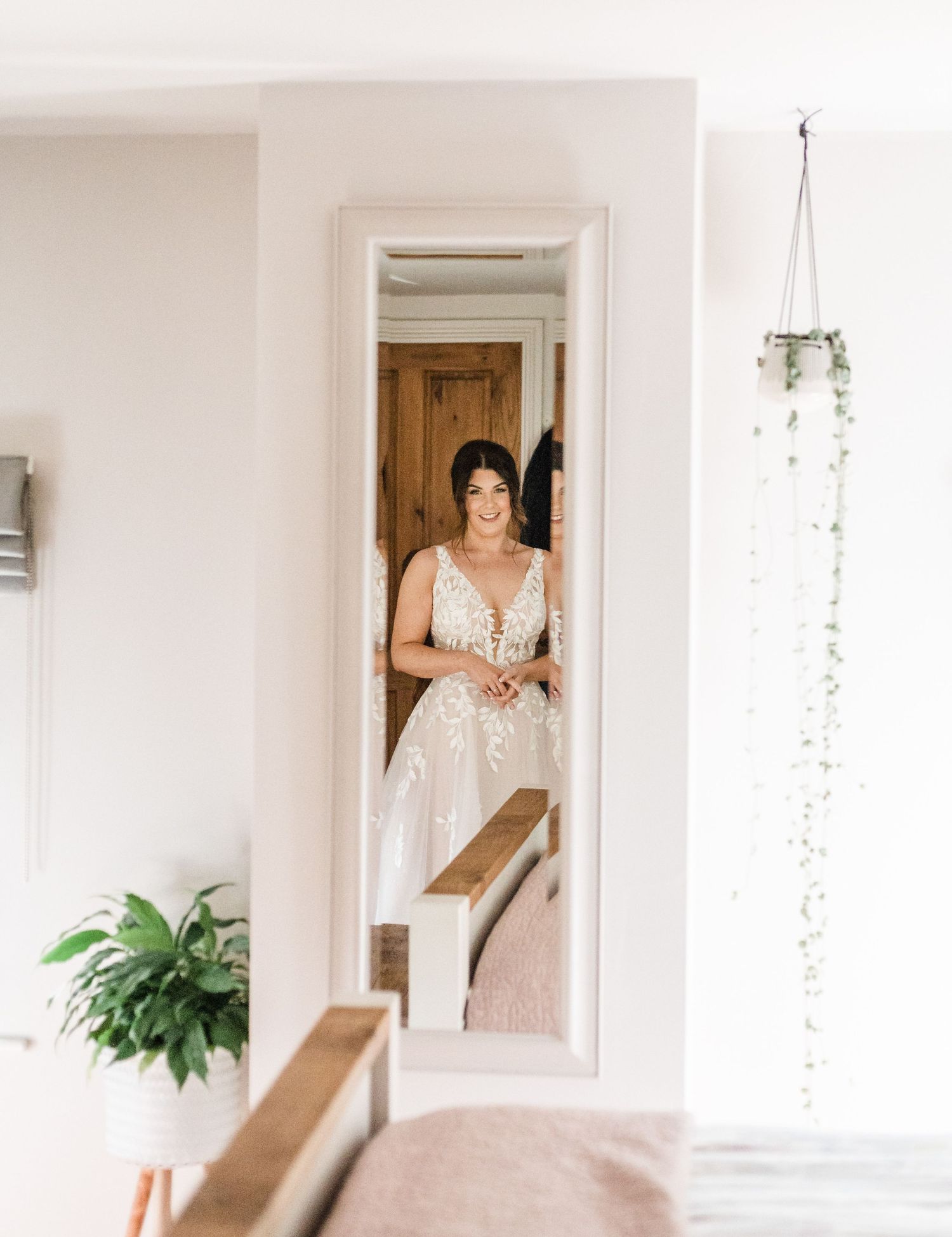 Beautiful white lace wedding dress reflected in a full-length mirror with hanging plants and natural light.