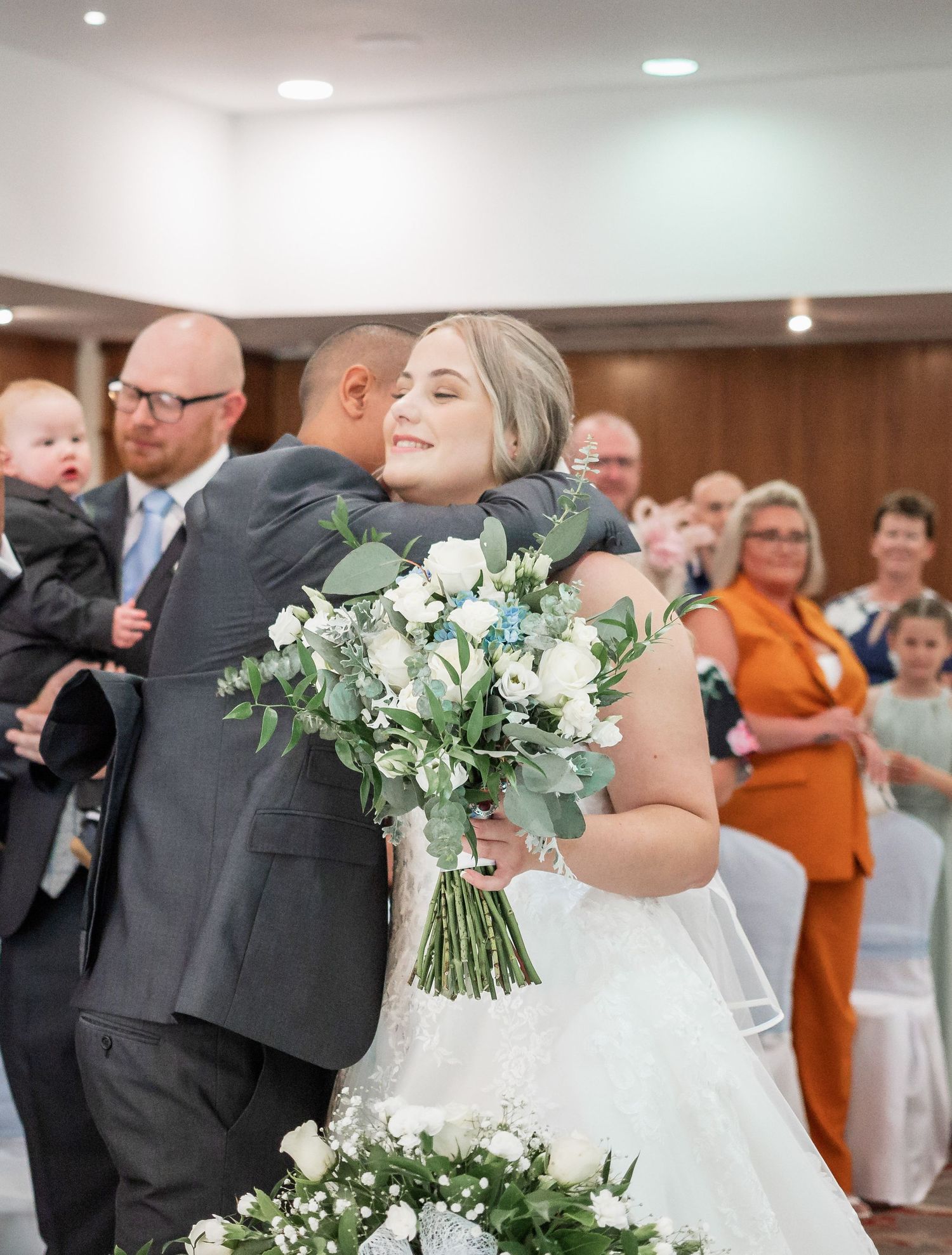 Bride with white bouquet embraces guest during wedding ceremony.