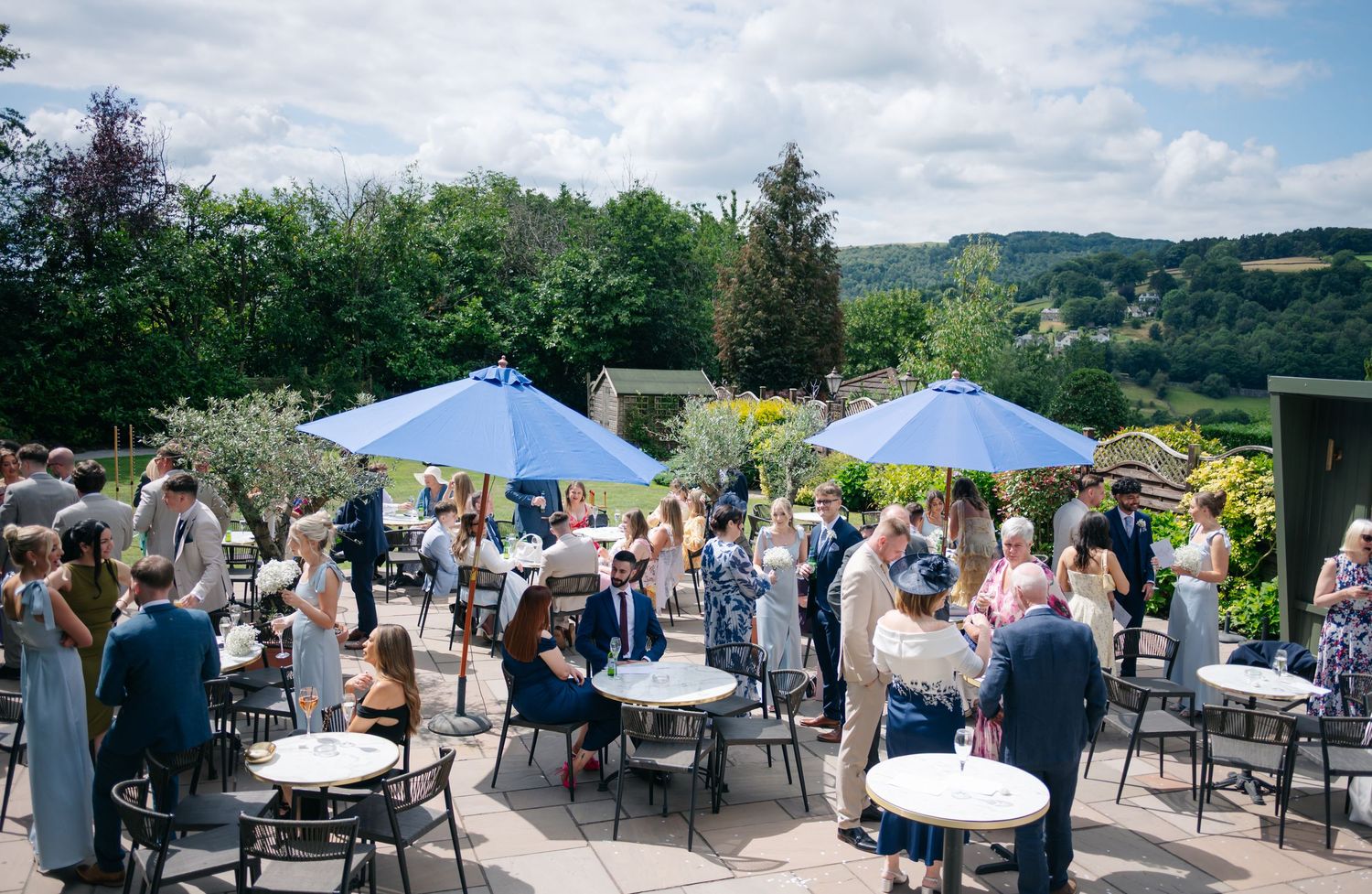 Panoramic view of an outdoor wedding reception with blue umbrellas and mountain scenery.