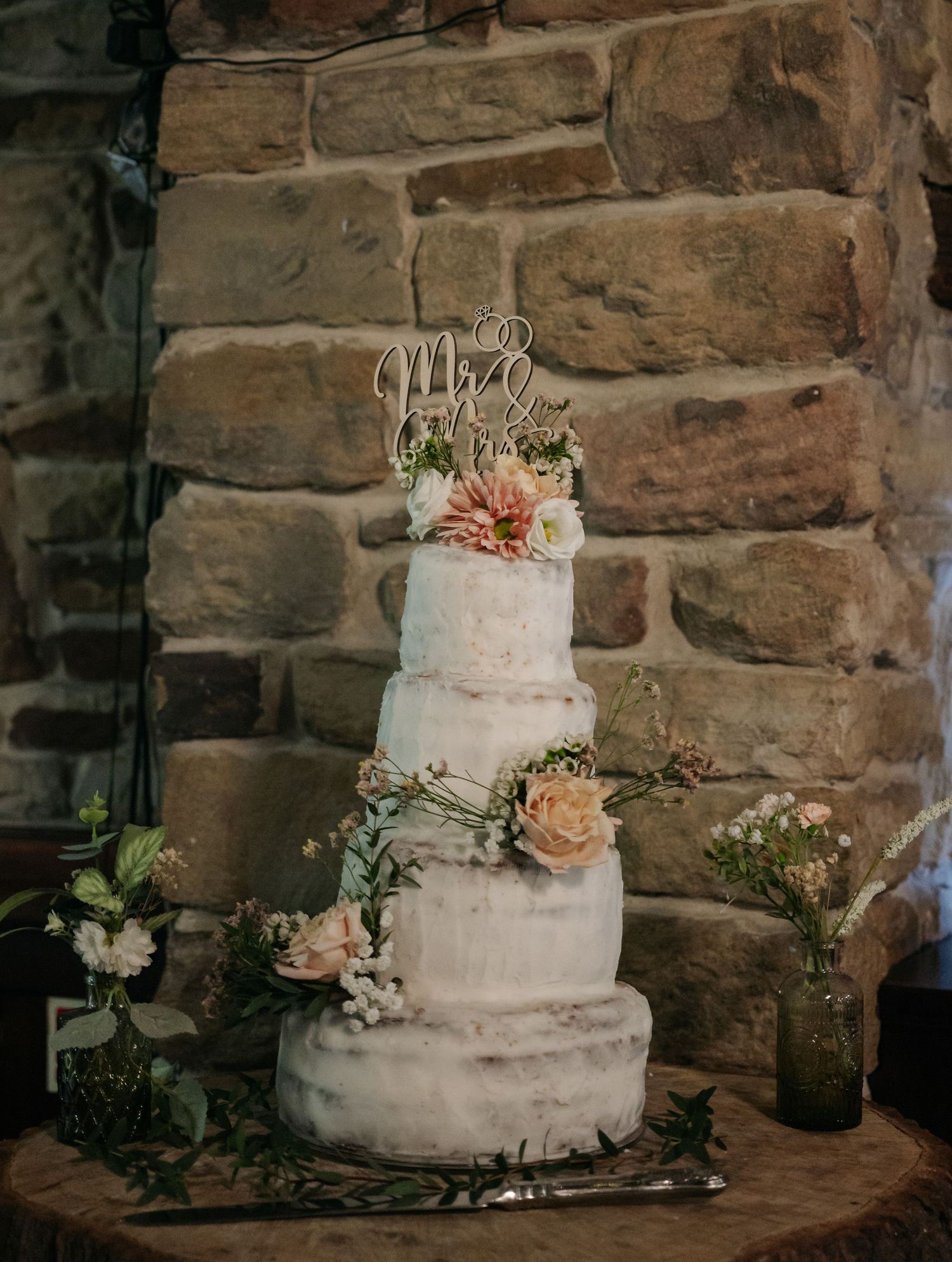An elegant three-tiered white wedding cake decorated with peach roses against a rustic stone wall.