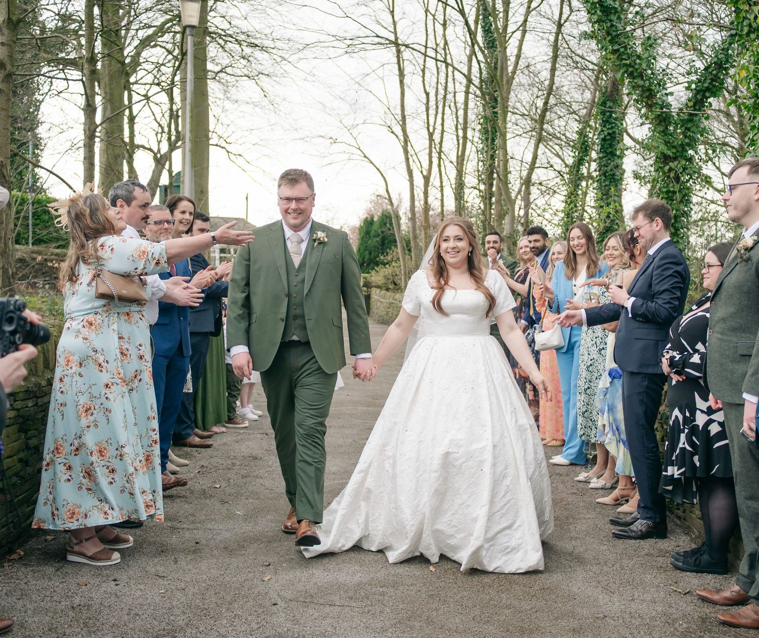 Wedding party and guests lined up outdoors for photos along a tree-lined path.