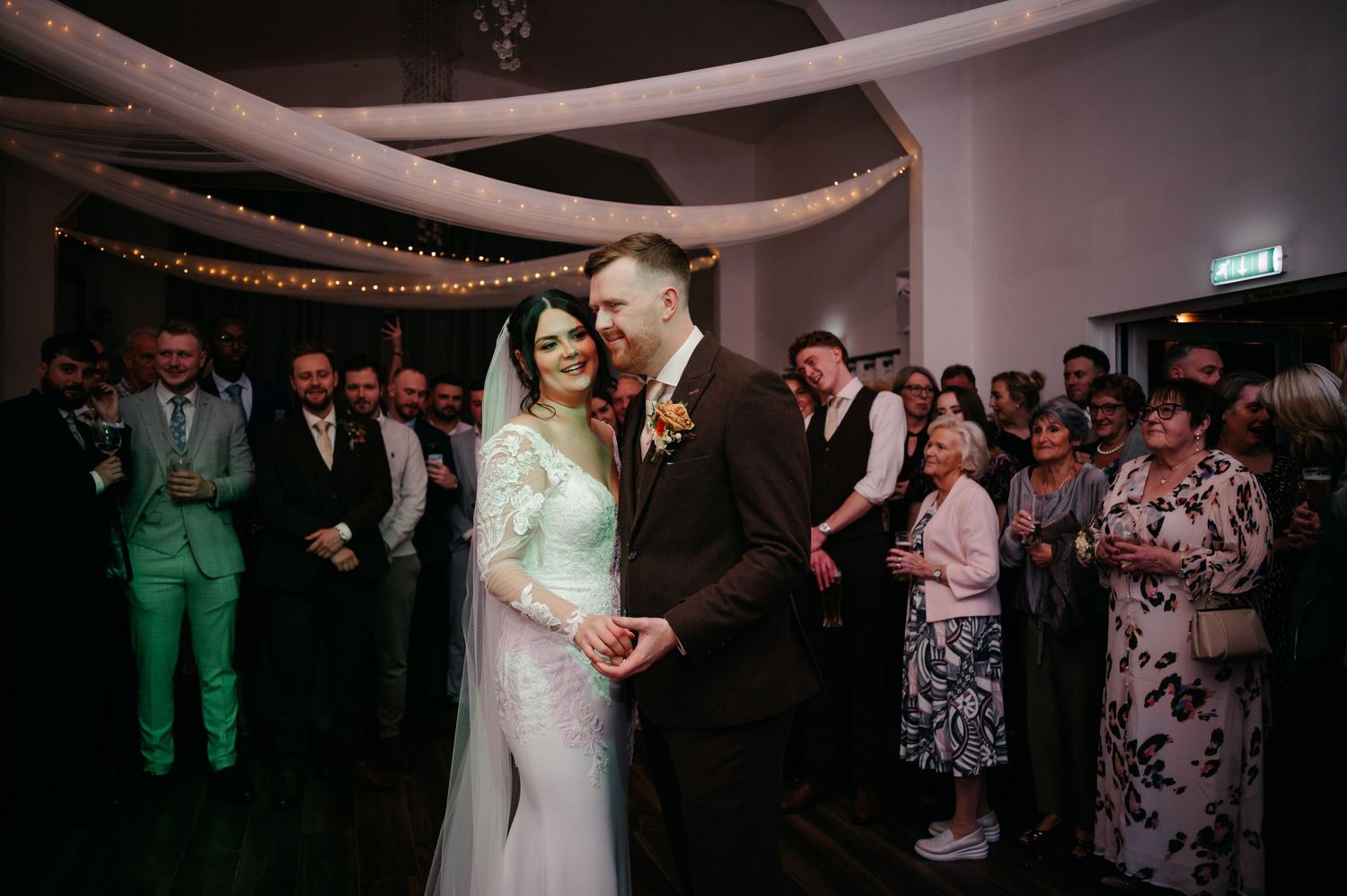 Wedding party dancing under romantic lighting and white fabric draping at evening reception.