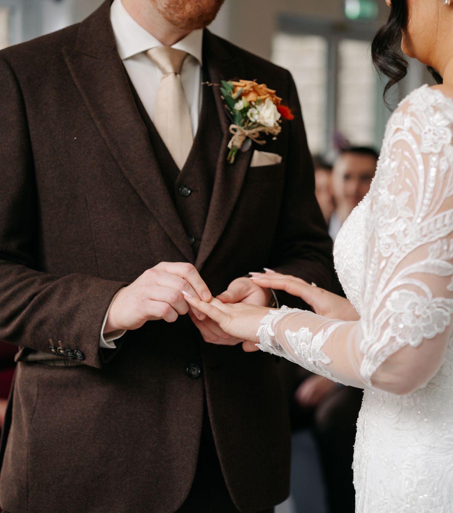 Close-up of wedding ring exchange with groom in brown suit and bride in lace sleeve dress.