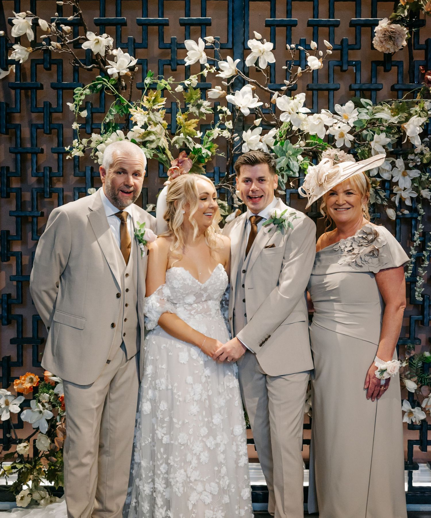 Wedding party poses together against a floral wall wearing light gray suits and a lace gown.