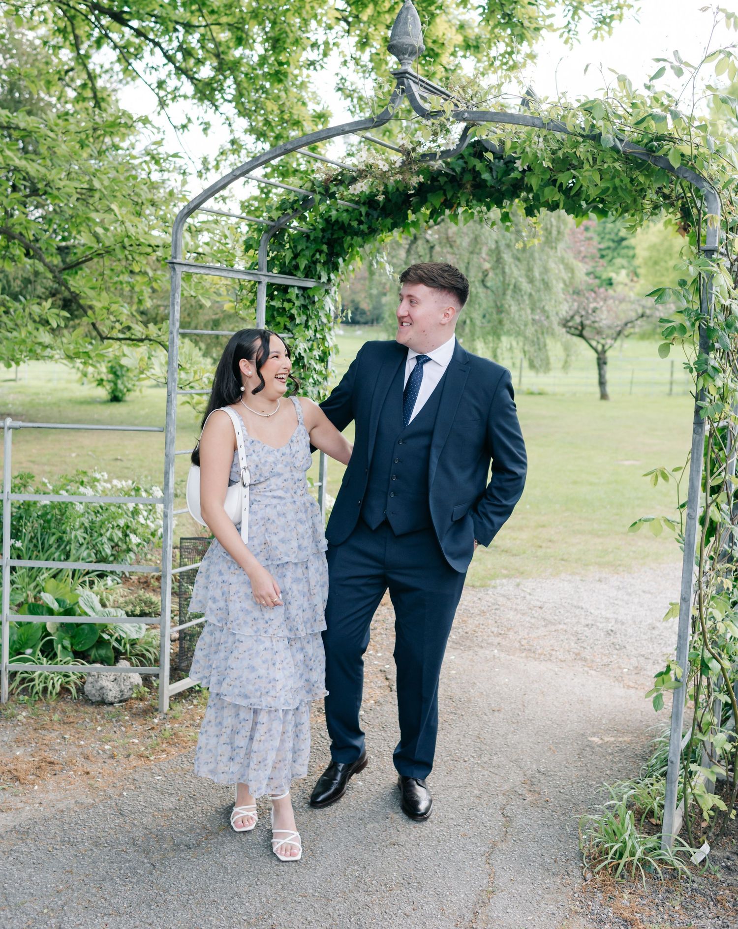 A couple walks together under a garden archway covered in greenery during a photo session.
