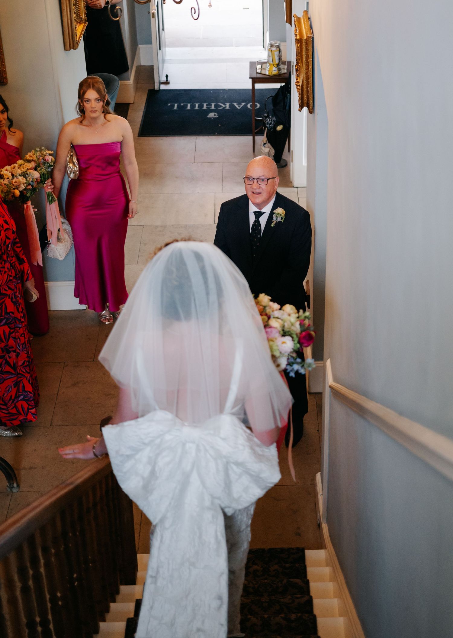 A bride in a white wedding dress descends a staircase while others watch from below.