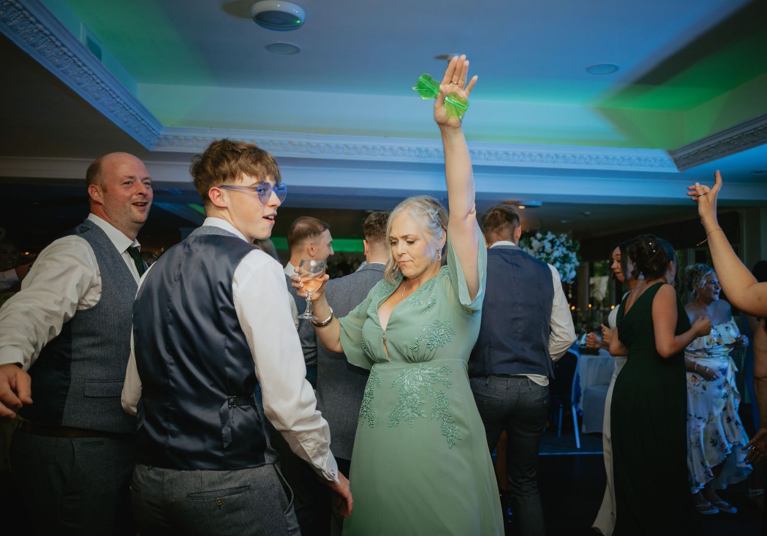 Wedding guests dance and celebrate on illuminated dance floor with blue and green lighting.