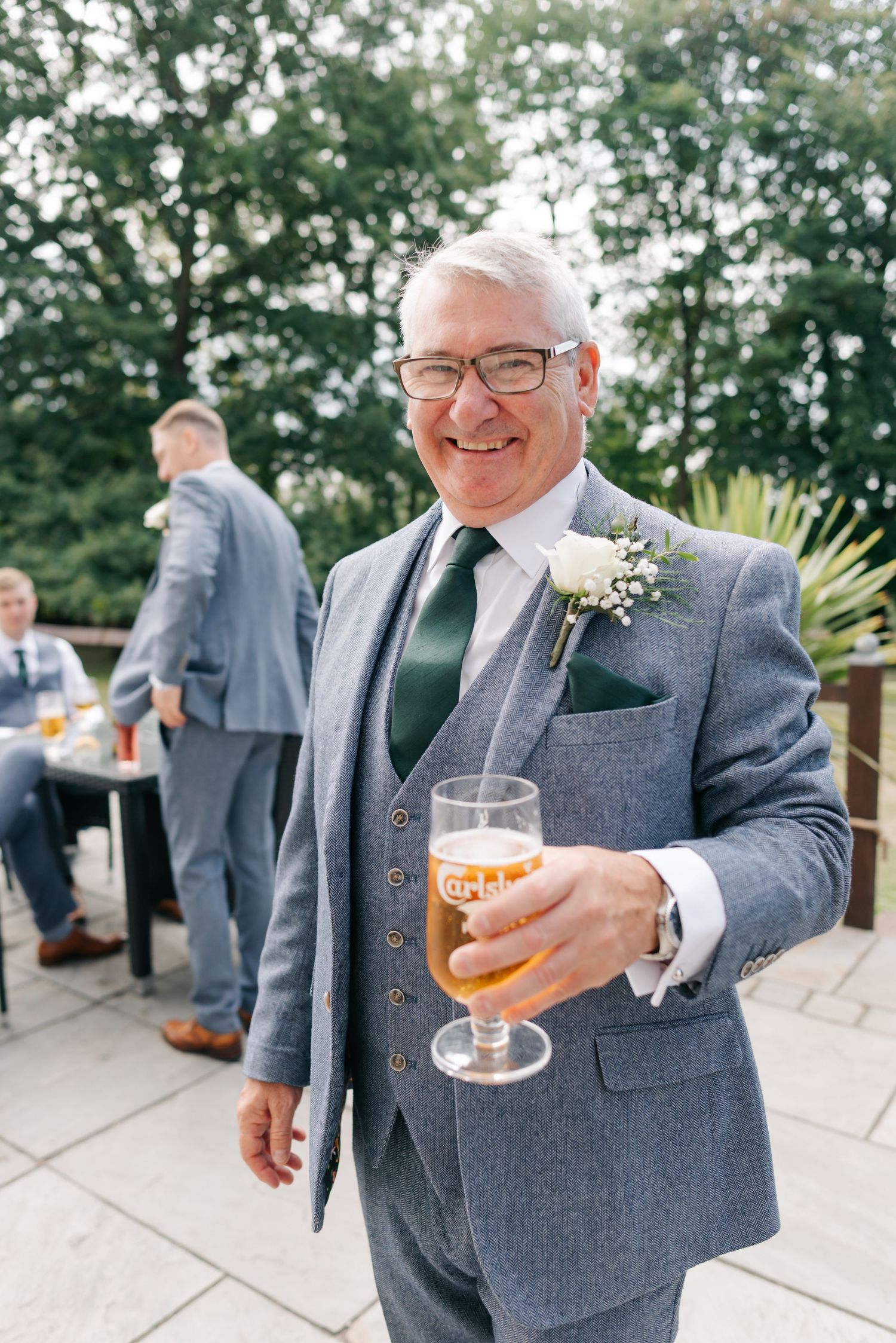 Groomsmen in gray suits share drinks and laughs on an outdoor patio during a wedding celebration.