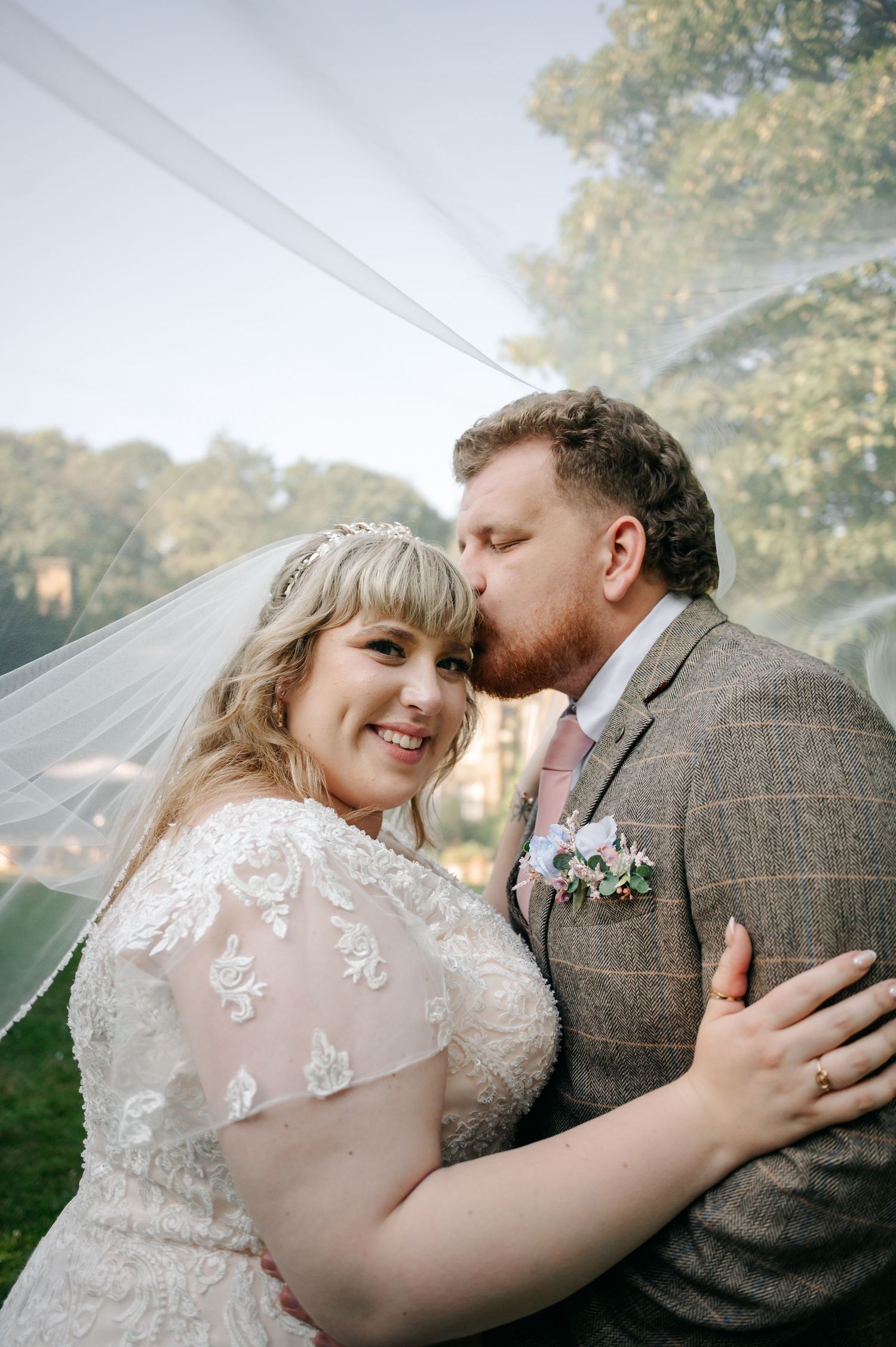 Groom in tweed suit kissing bride on the forehead under a flowing veil in an outdoor garden setting.
