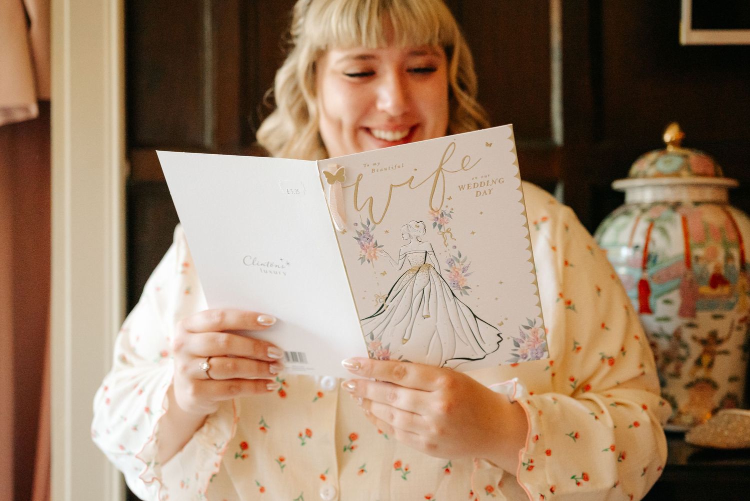 Smiling blonde woman reading a Wife wedding card with floral dress illustration, holding two cards indoors.