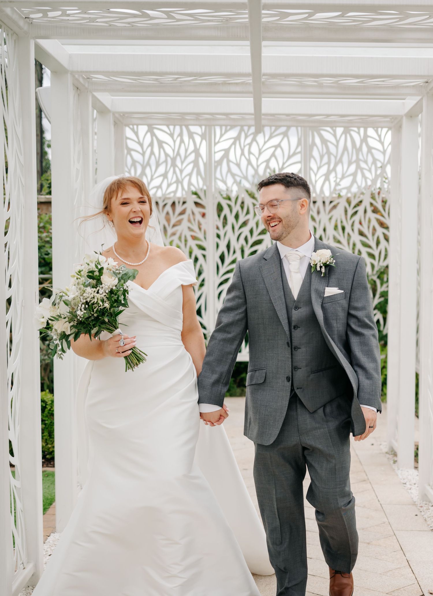 A couple shares joyful laughter while walking together through an elegant white pergola walkway decorated with greenery.