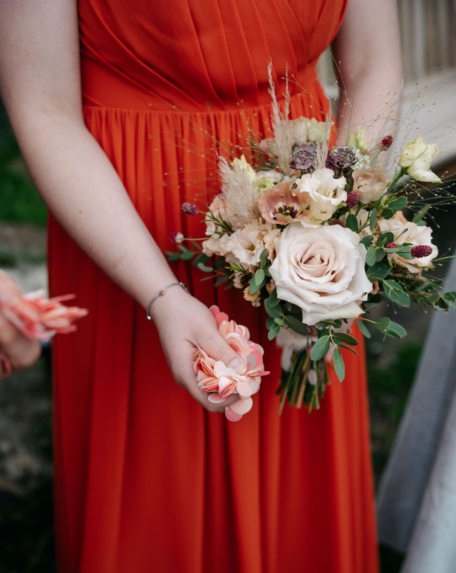 Bridesmaid in a coral red chiffon dress holding a wedding bouquet of cream roses, peach blooms and dried grasses.