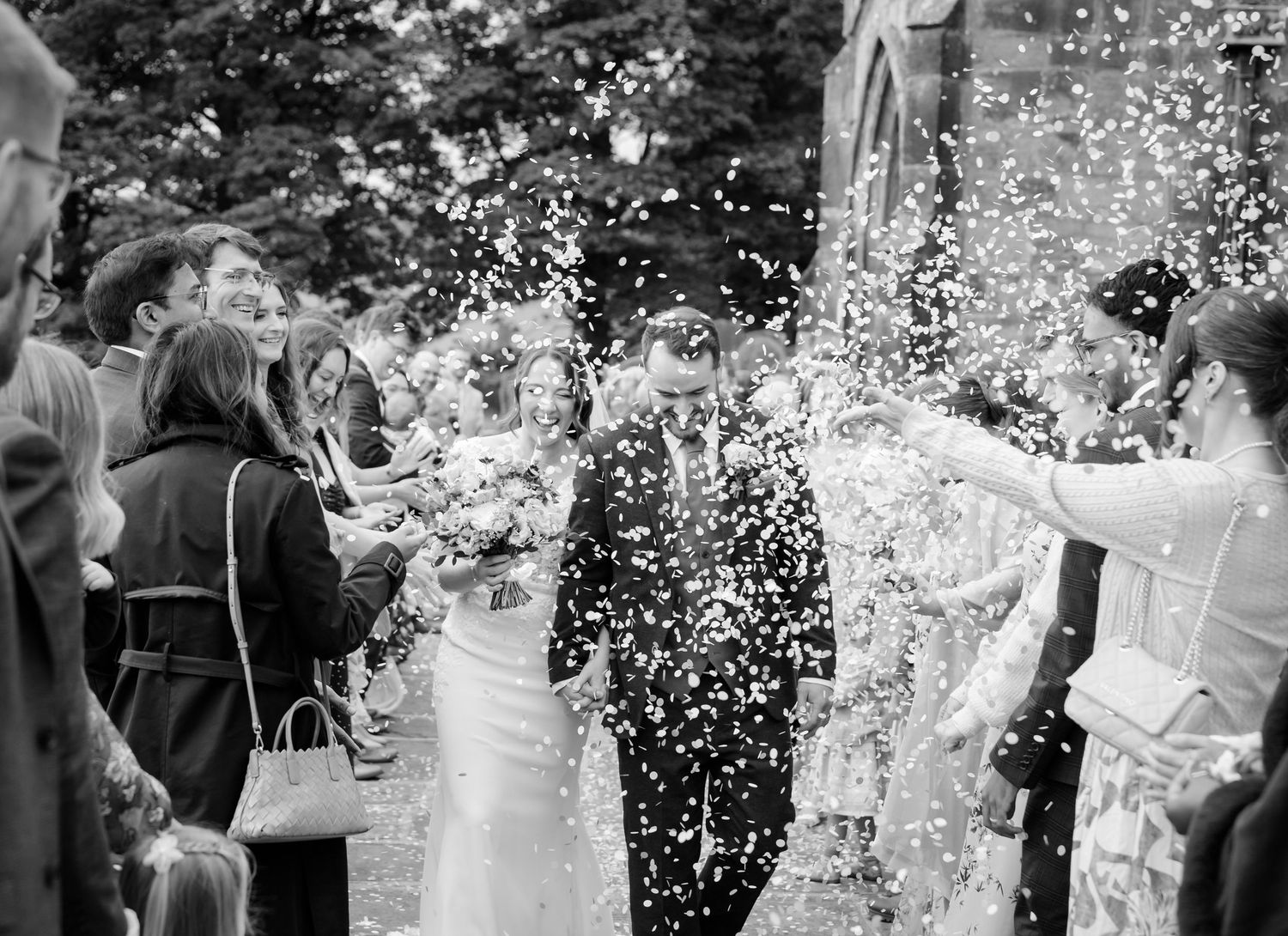Black and white wedding photo of a bride and groom walking through guests tossing confetti outside a stone church.