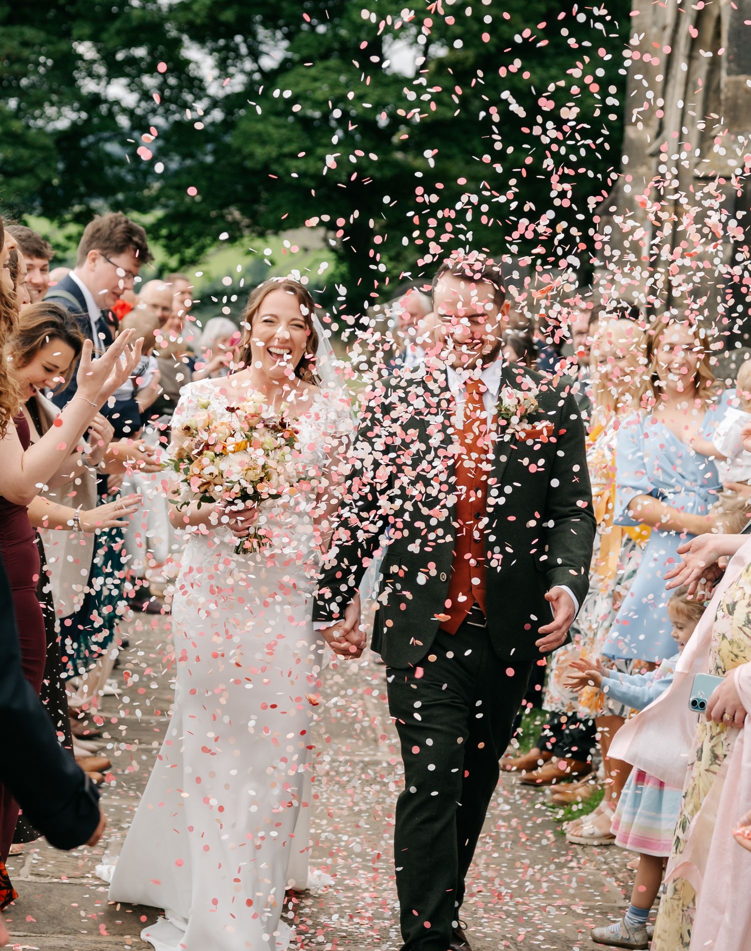 Smiling bride and groom hold hands as wedding guests shower them with pink confetti petals outside the ceremony.