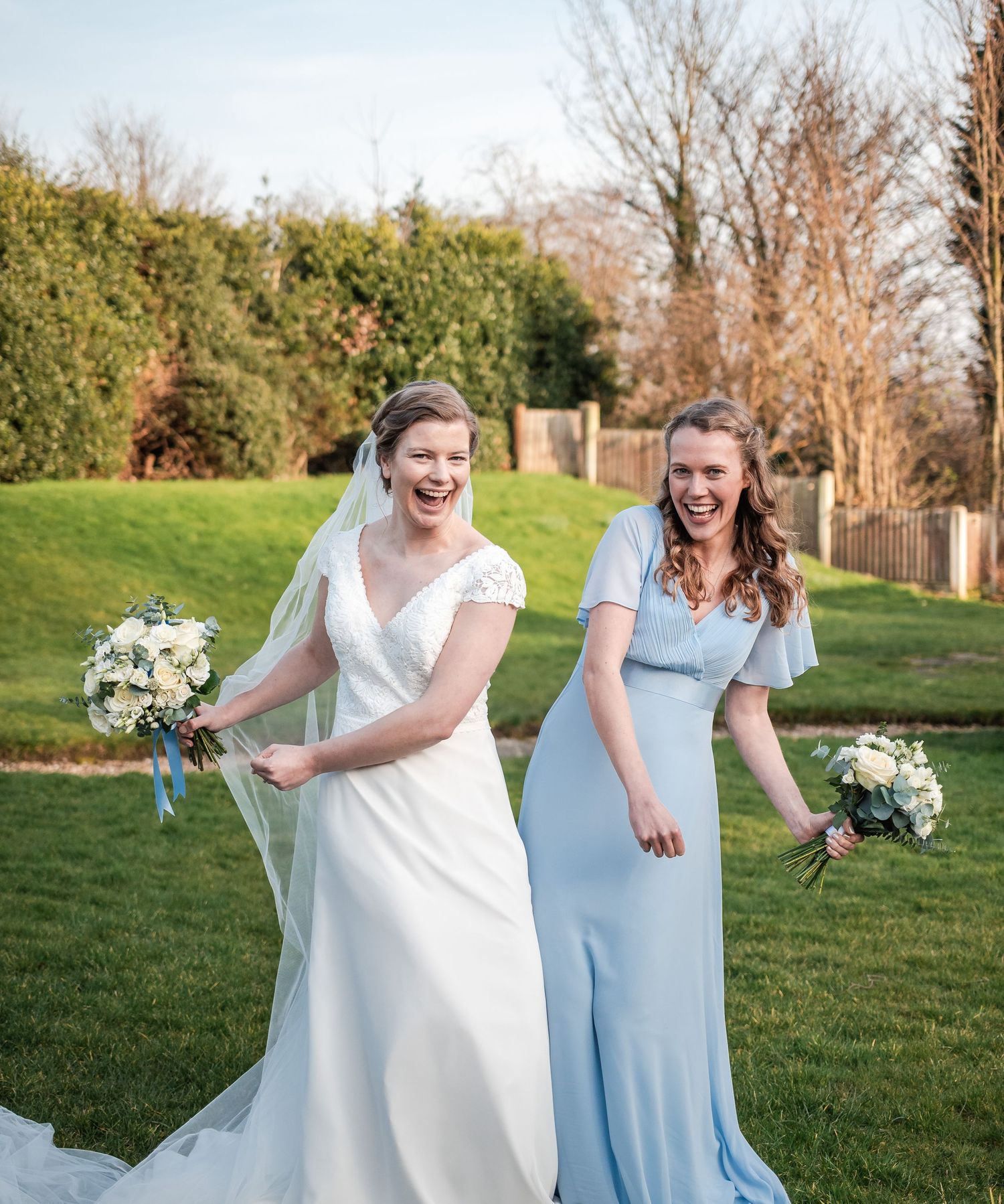 Bridal party in white and light blue dresses laughing together on a grassy hill.