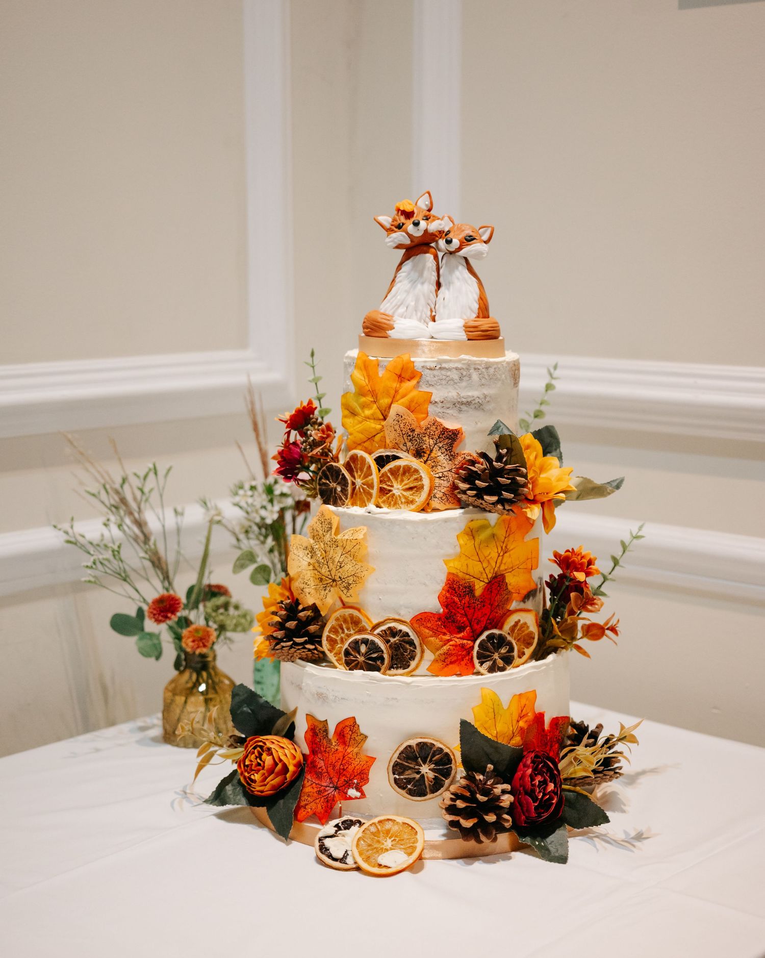 Three-tiered autumn wedding cake decorated with orange flowers, pinecones, and dried citrus slices.