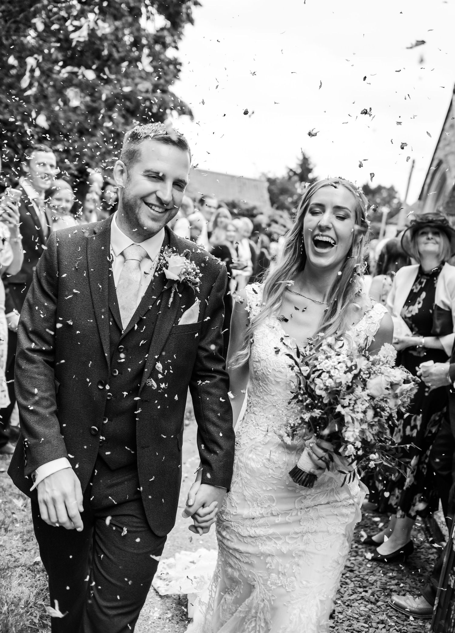 Joyful newlyweds walk through shower of white confetti after outdoor wedding ceremony.