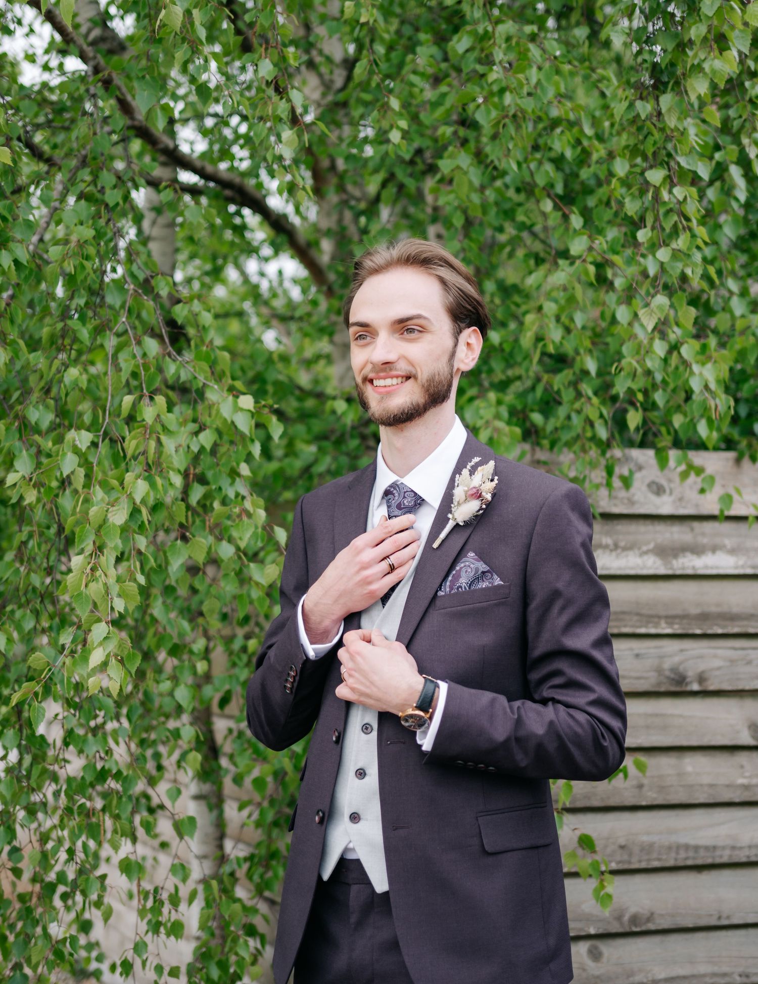 Wedding portrait of person in formal black tuxedo and vest against green foliage background.