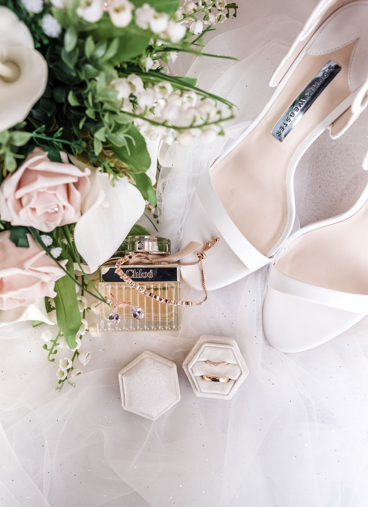Elegant white bridal shoes with pink roses and perfume bottle on white surface.