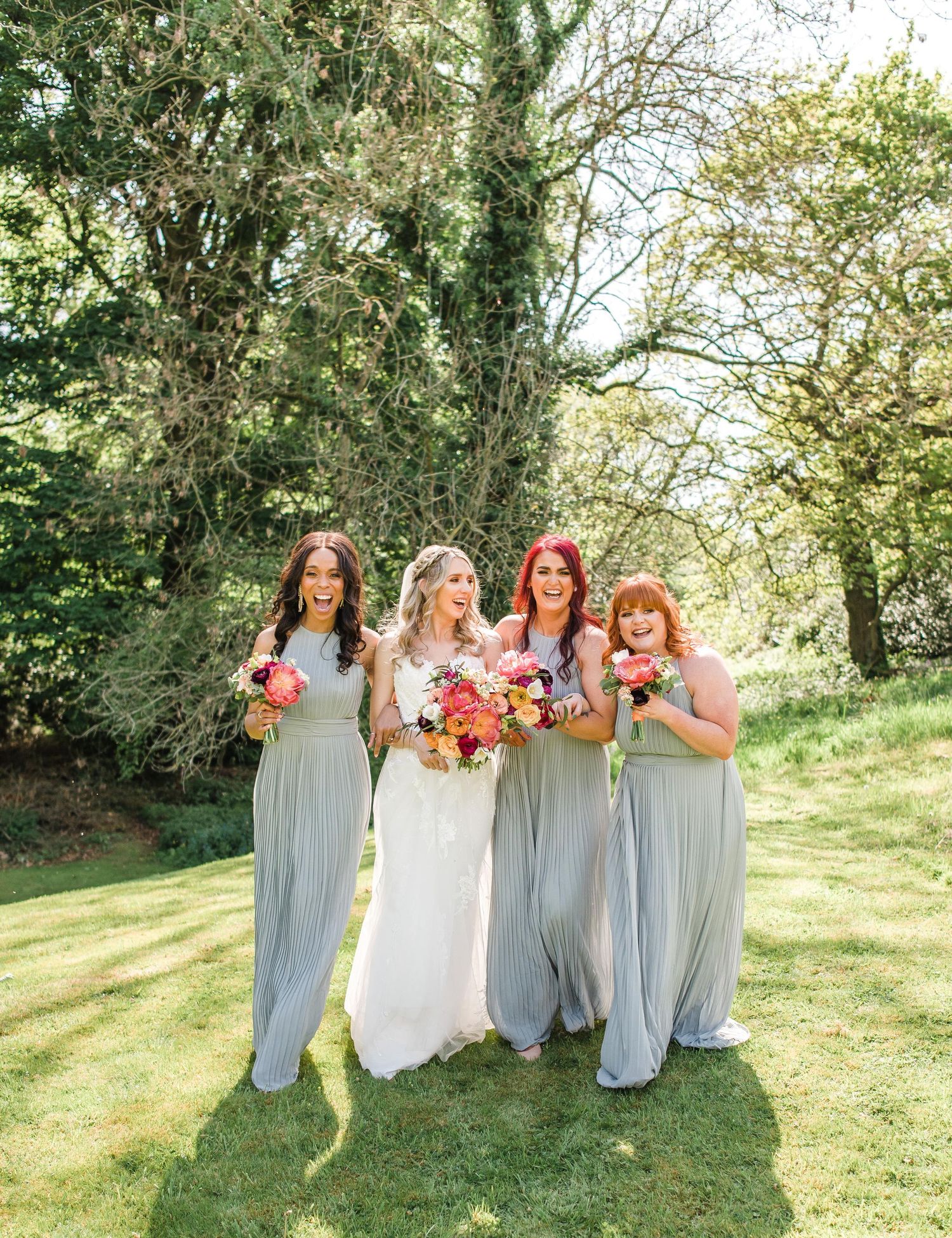 Bridal party in flowing sage green dresses pose with vibrant bouquets in sunny garden setting.