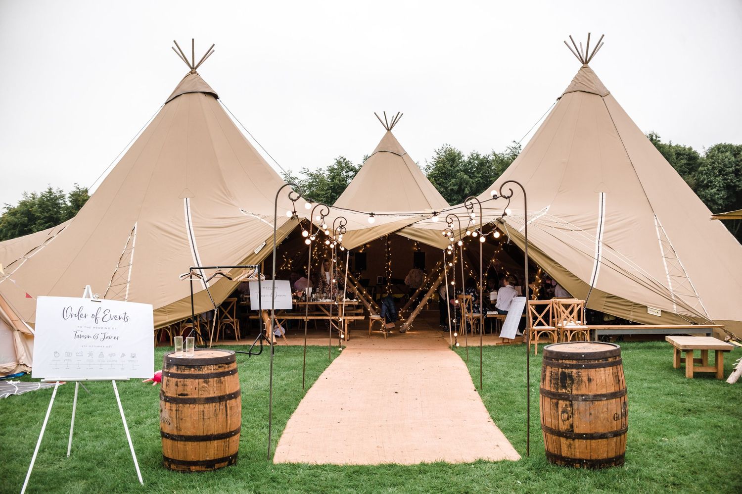Three connected tipi-style wedding tents set up on grass with wooden barrels and decorative pathway.