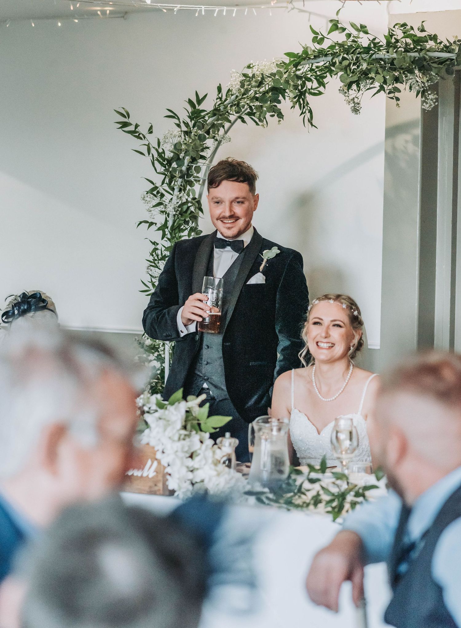 Newlyweds stand for toast at wedding reception under decorative greenery arch.